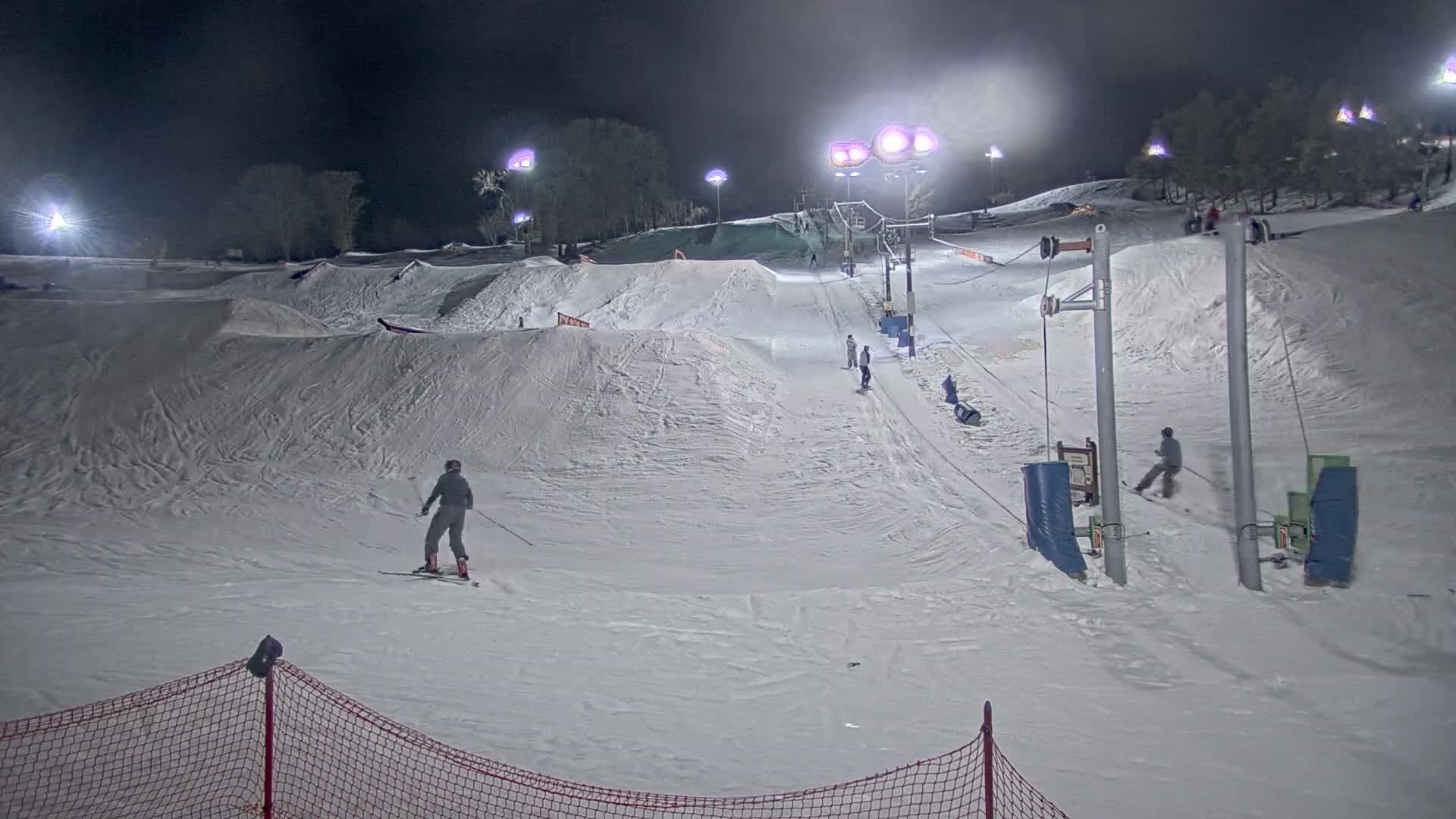 A nighttime view of a snow-covered ski slope with several skiers and snowboarders, illuminated by artificial lights, features terrain park features and a surface lift.