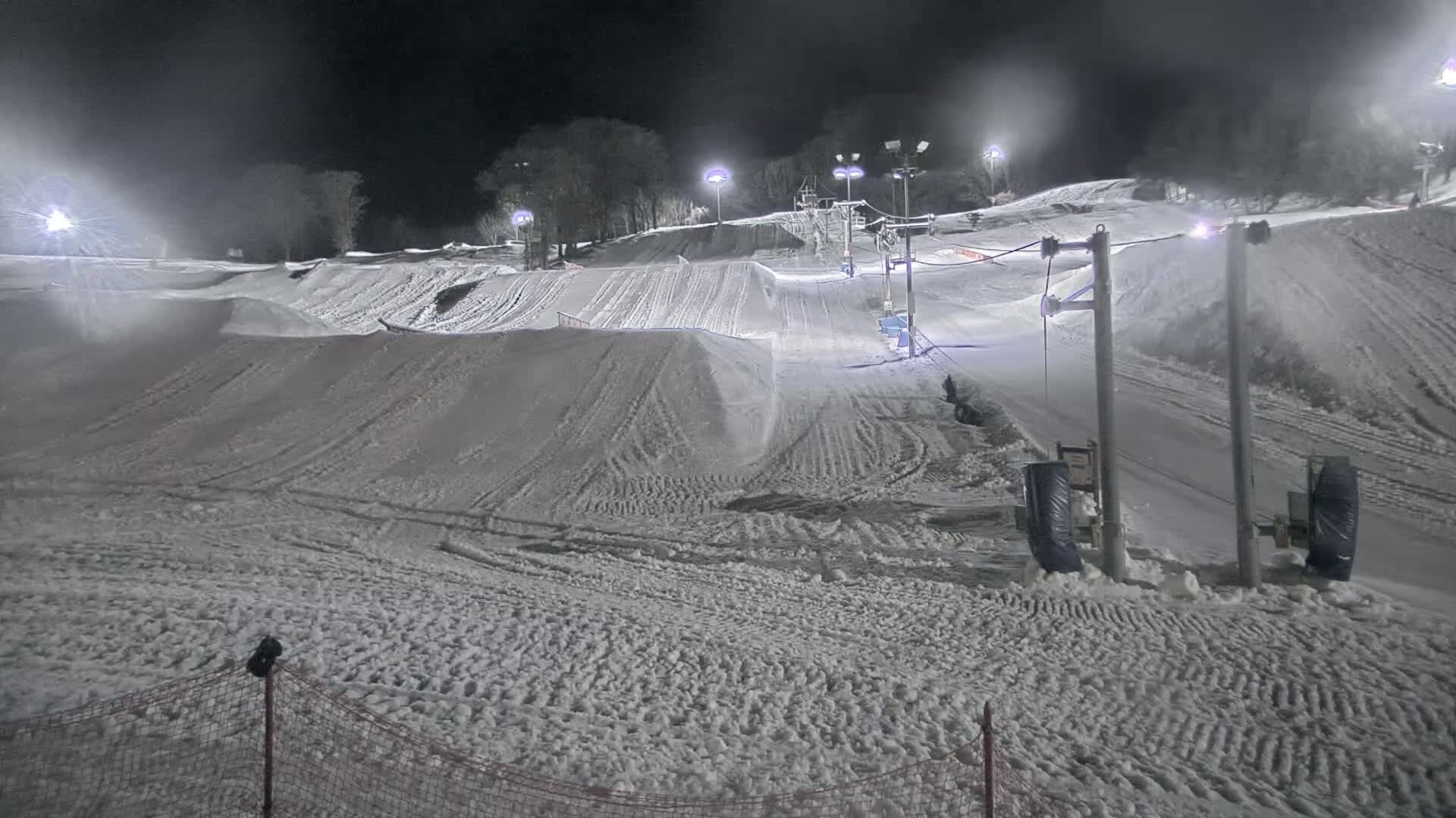 A snow-covered ski slope at night, illuminated by artificial lights, features a ski lift and various snow jumps.