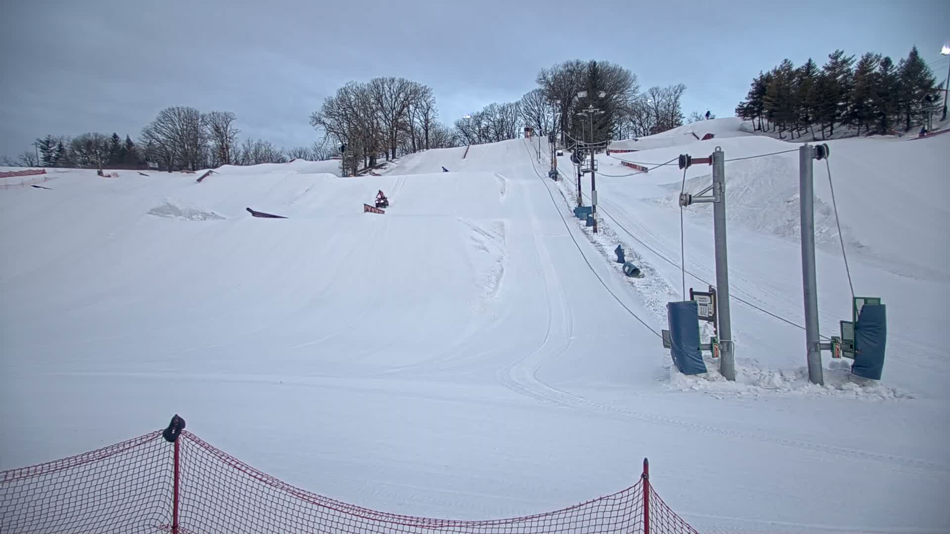 A snow-covered ski slope with a surface lift and a few skiers or snowboarders visible under an overcast sky.