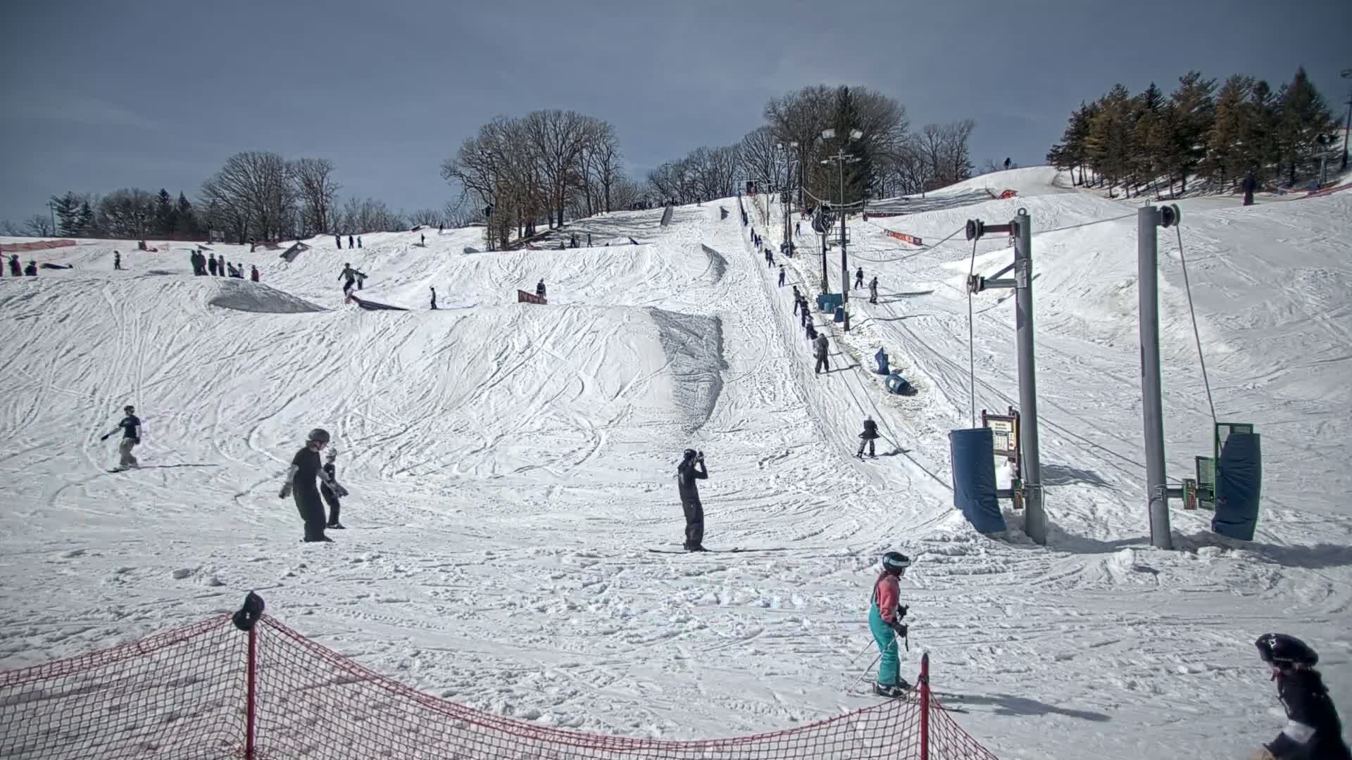 A sunny day at a snow-covered ski slope shows skiers and snowboarders of various skill levels enjoying the terrain, with a ski lift visible in the background.
