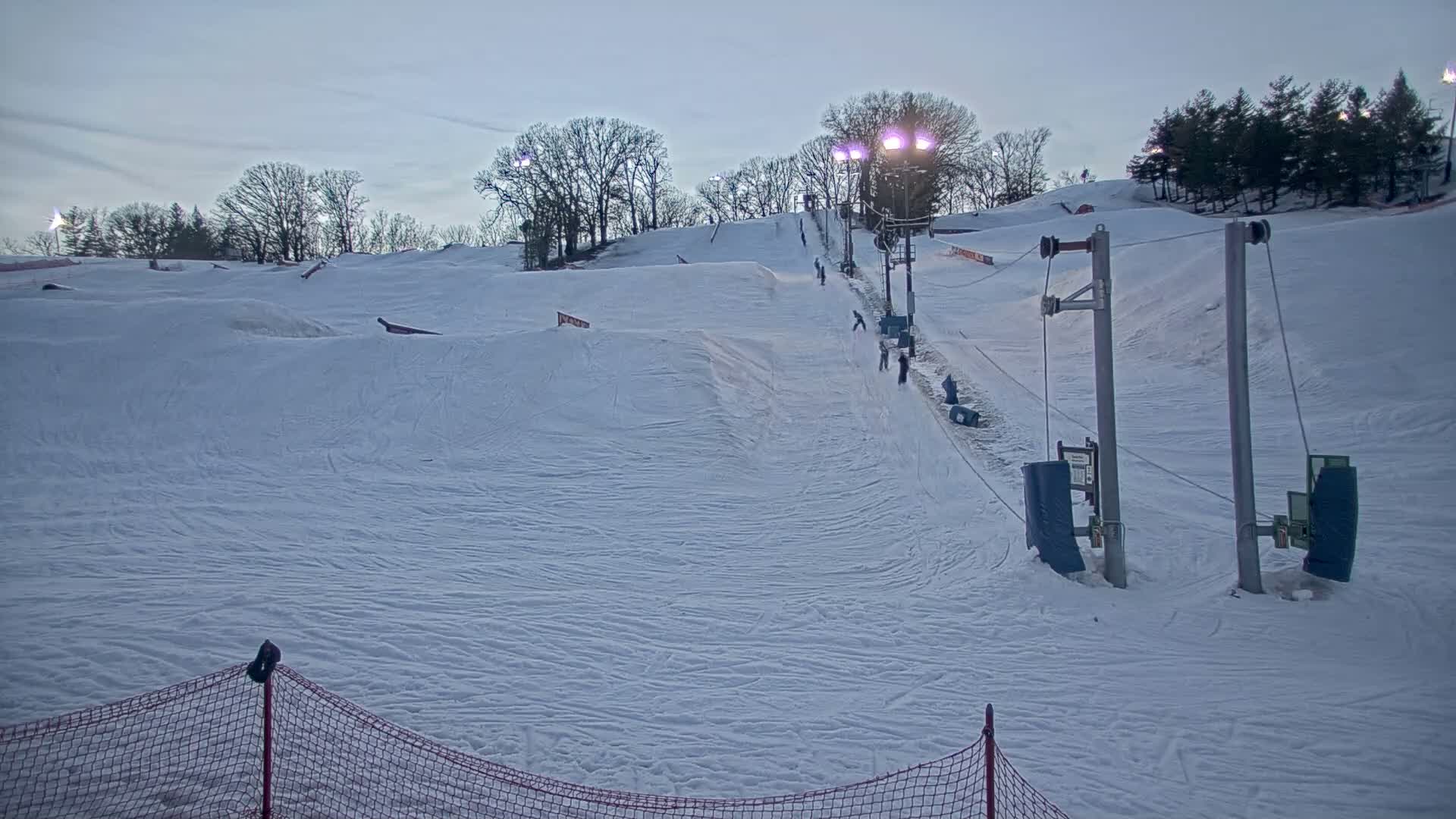 A snow-covered ski slope with several people skiing or snowboarding down under a twilight sky.