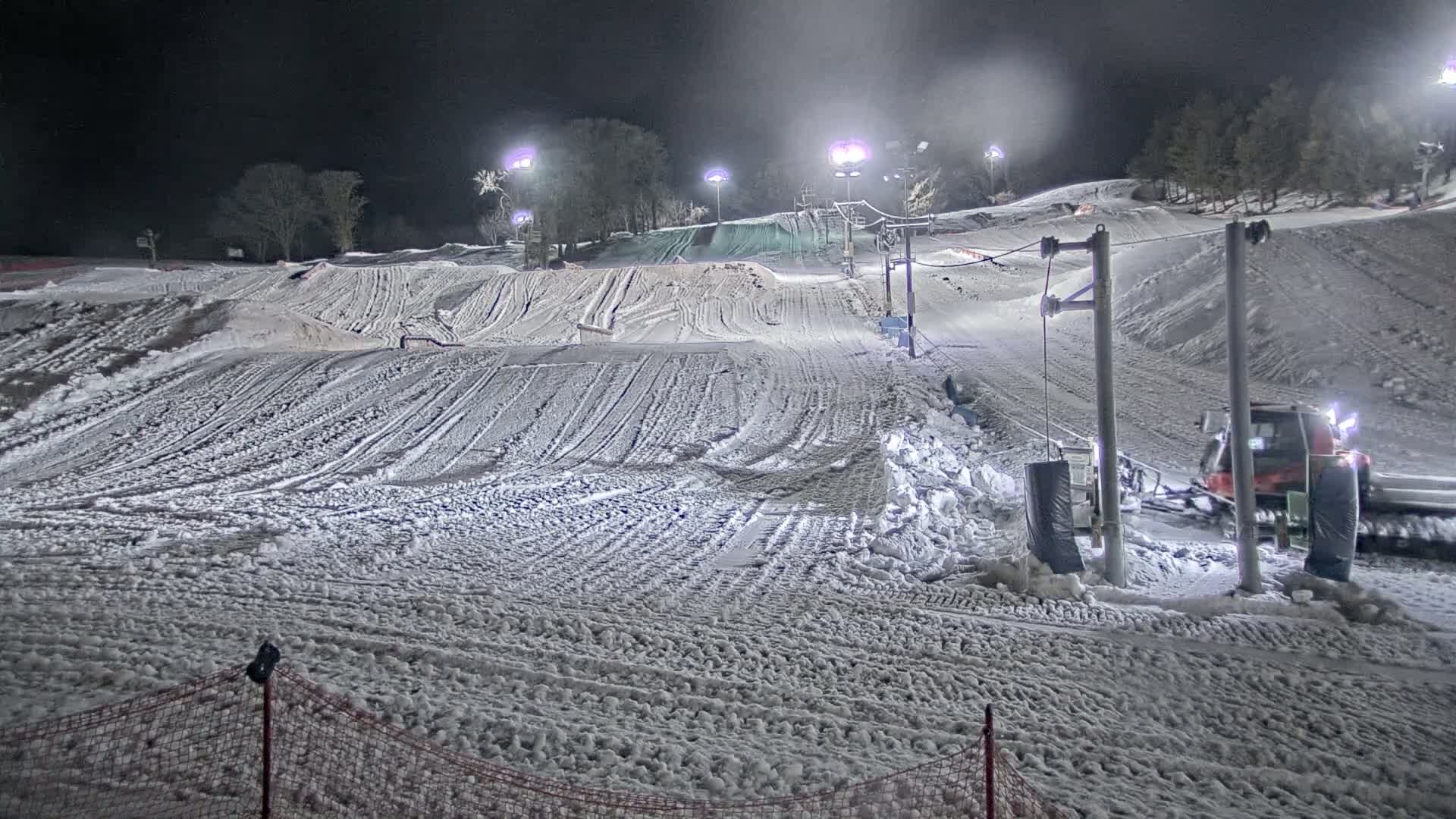 A snow-covered ski slope at night is illuminated by artificial lights, with a snow groomer visible near a ski lift.