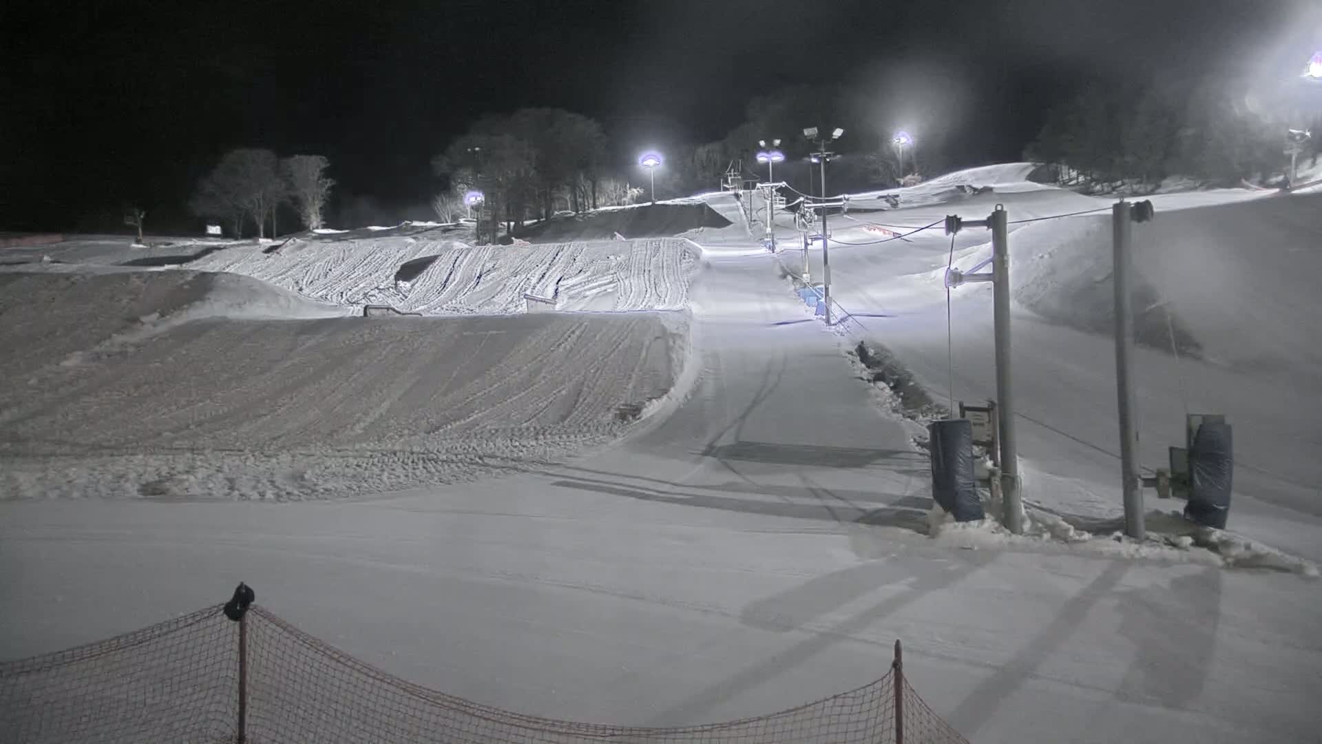 A snow-covered ski slope at night, illuminated by artificial lights, features various runs and a ski lift.