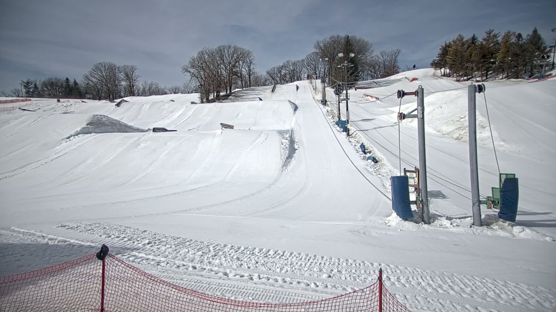 A snow-covered ski slope with terrain features and a ski lift under a partly cloudy sky.