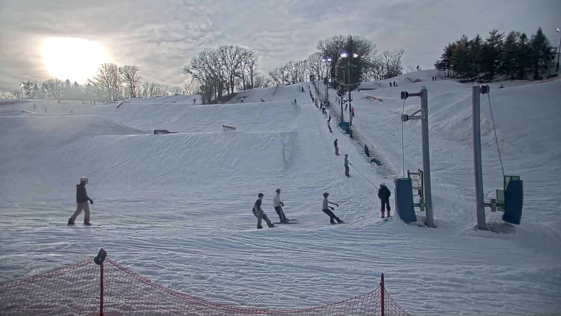 A snowy ski hill with several people snowboarding and skiing under a mostly cloudy sky.