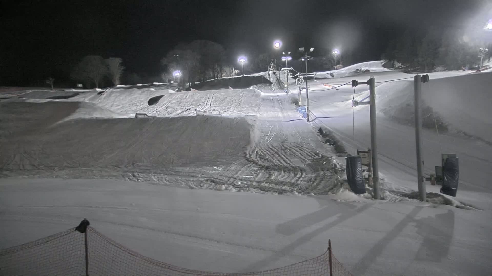 A snow-covered ski slope is illuminated at night by artificial lights, featuring a ski lift and various terrain features.