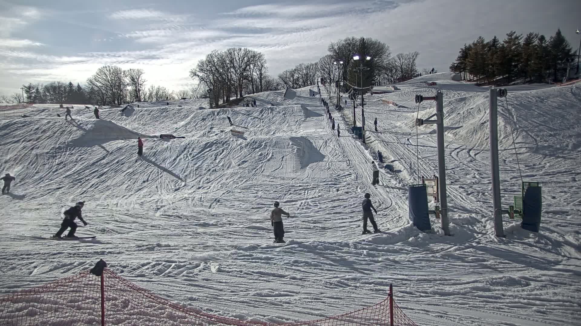 A snow-covered ski hill with several people snowboarding and skiing under a partly cloudy sky.