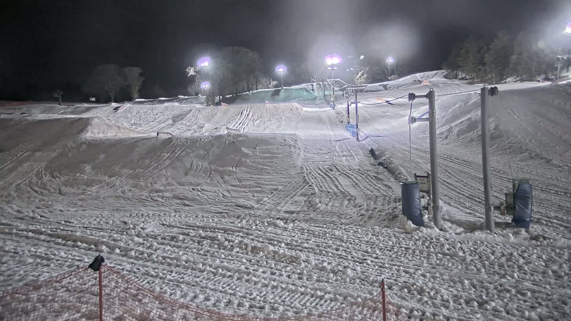 A snow-covered ski slope is illuminated at night by several lights, with a ski lift visible in the background and some snow terrain features.