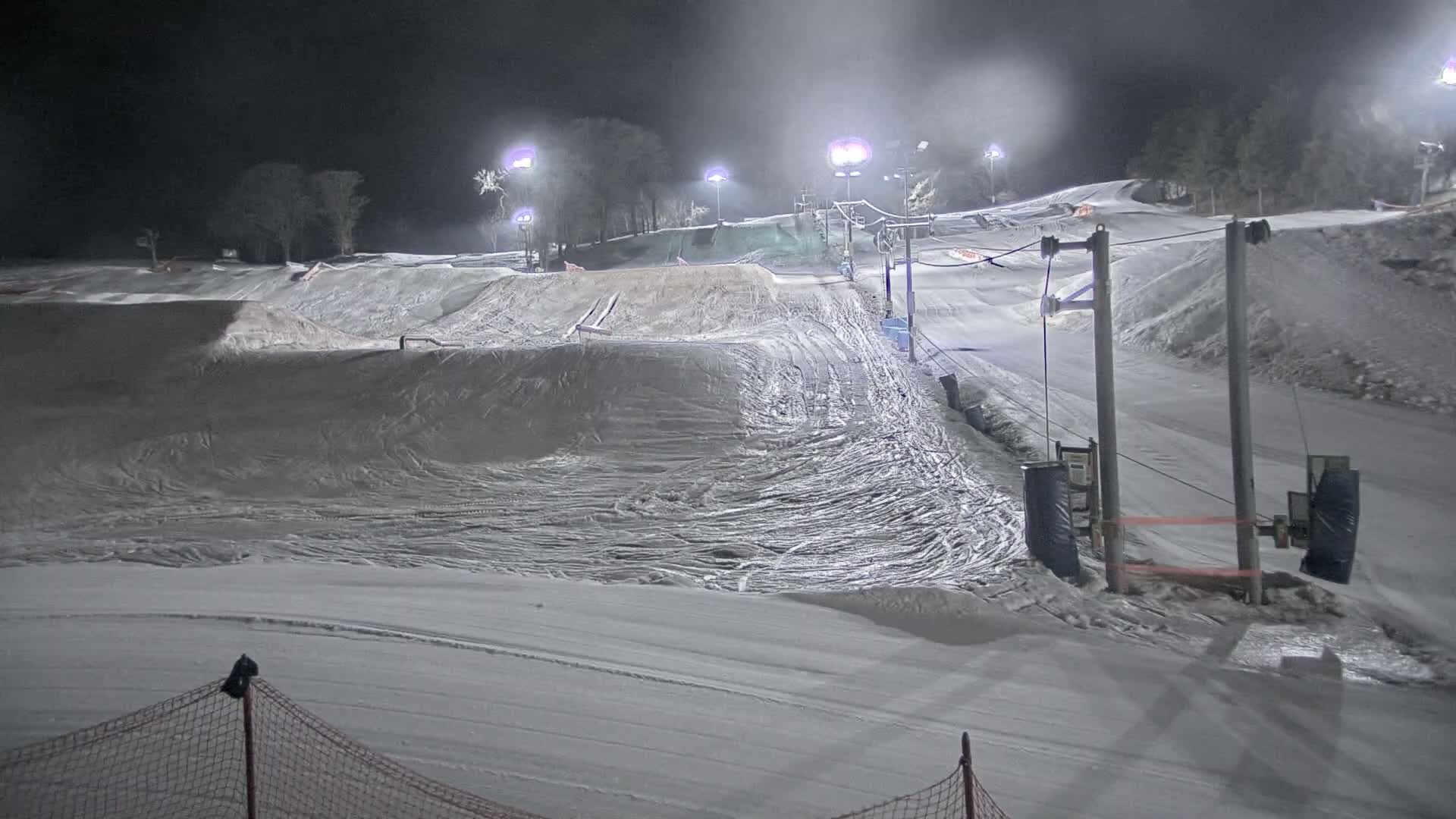 A snow-covered ski slope is illuminated at night by artificial lights, with some terrain features visible and a ski lift in the background.