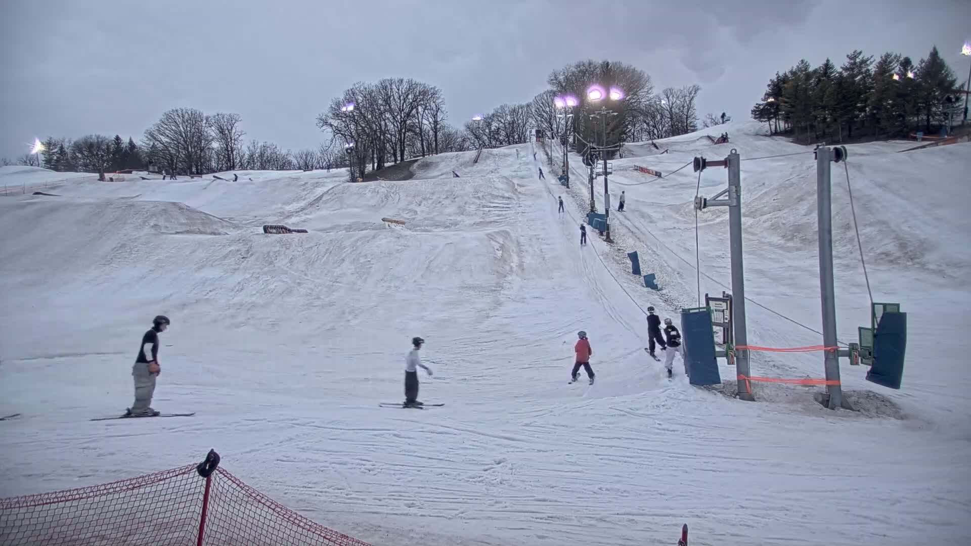 A snowy ski slope with several skiers and snowboarders, under an overcast sky.