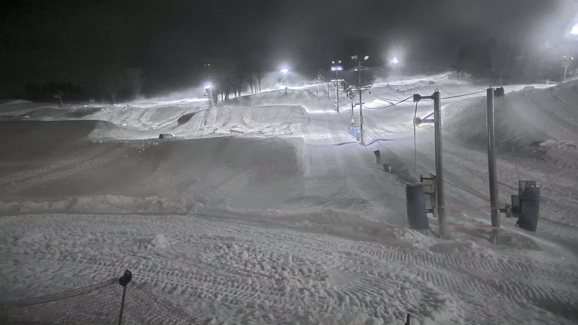A snow-covered terrain park with ski lifts is illuminated at night under snowy, windy conditions.