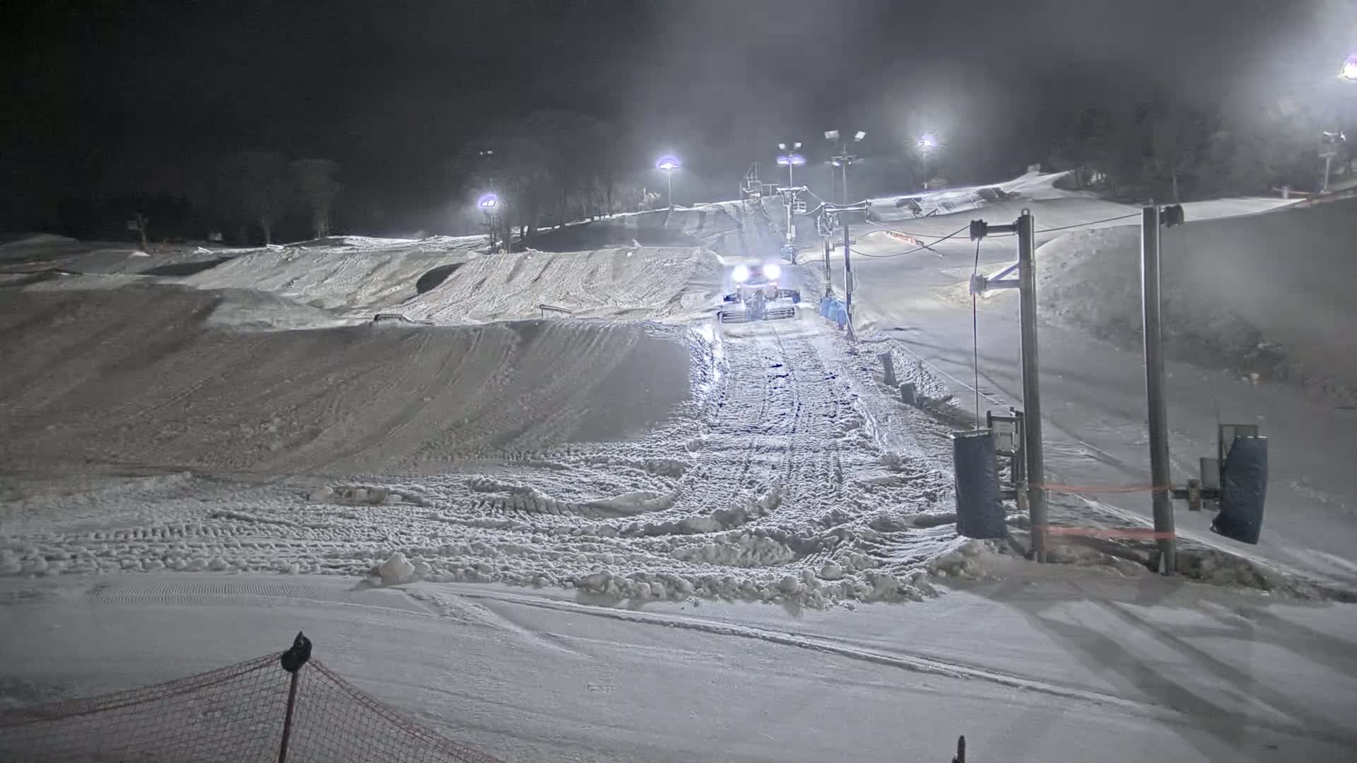 A snowcat grooms a snow-covered ski slope at night under artificial lights.