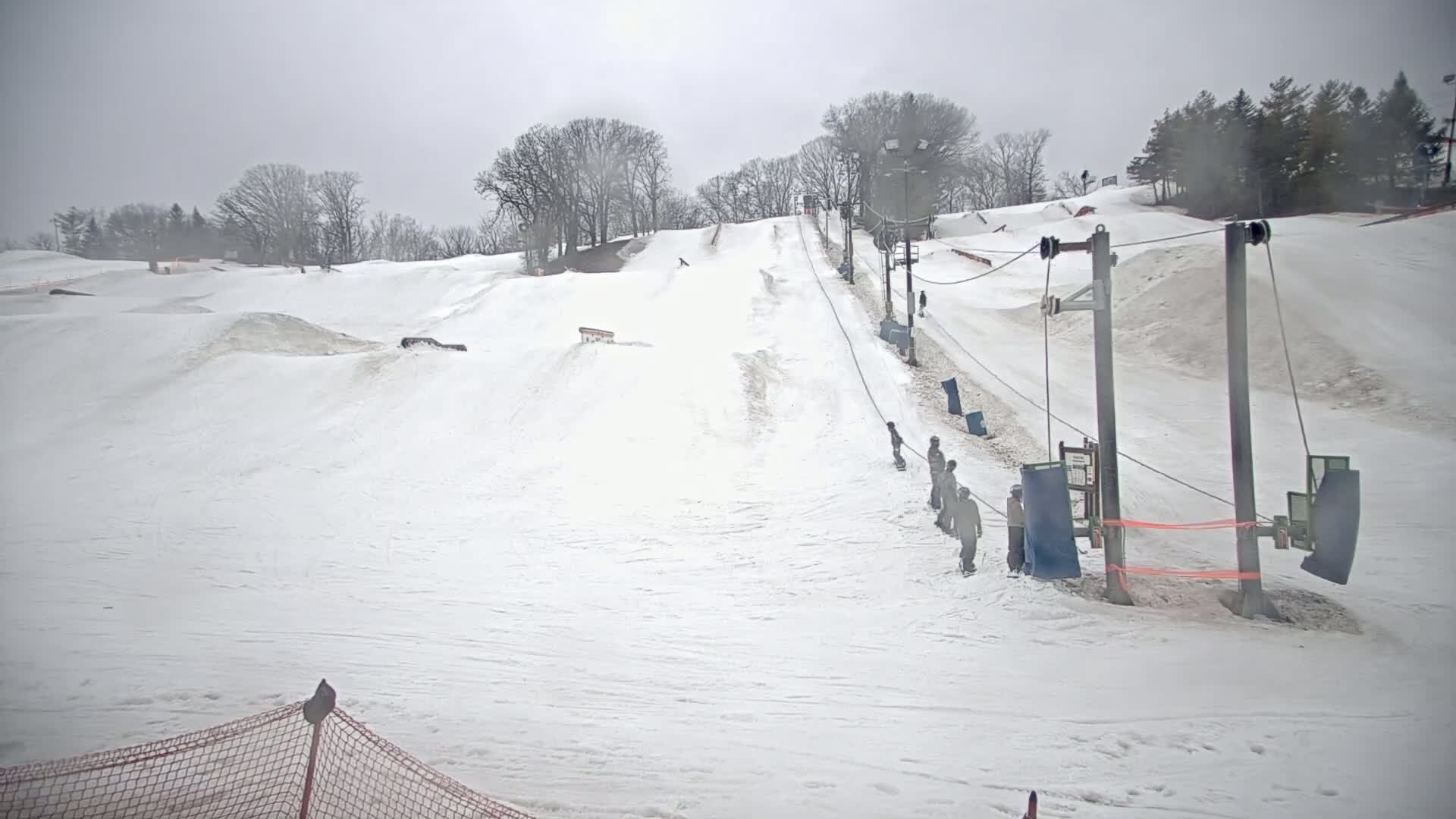 A snowy ski slope with a surface lift and several people snowboarding or skiing under an overcast sky.