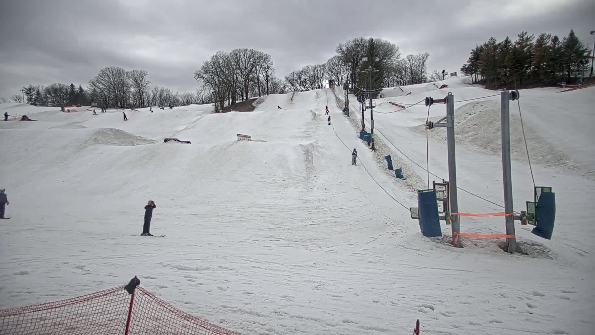 A snowy ski slope with a surface lift and several skiers and snowboarders under an overcast sky.