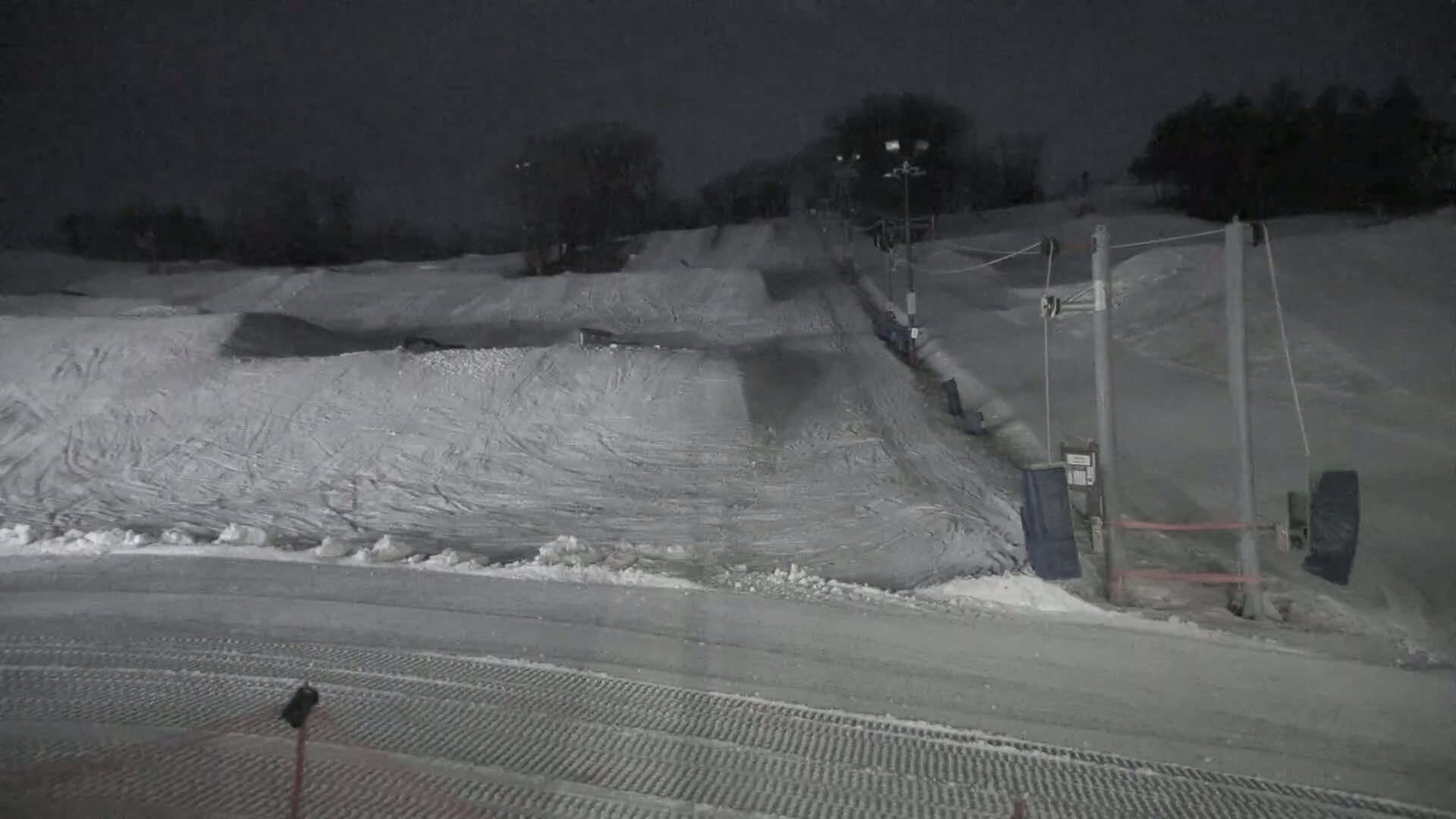 A snow-covered terrain park at night, illuminated by artificial lights, features various jumps and ramps, with a ski lift visible in the background.