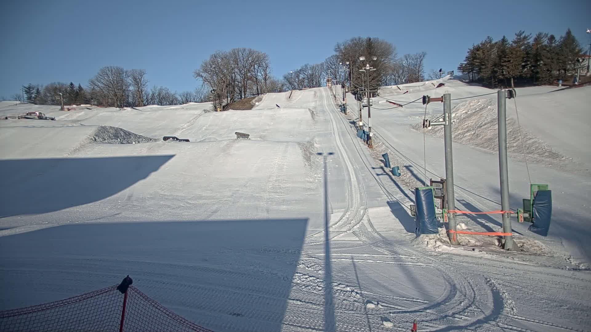 A snow-covered ski slope with a ski lift and surrounding trees under a clear, sunny sky.
