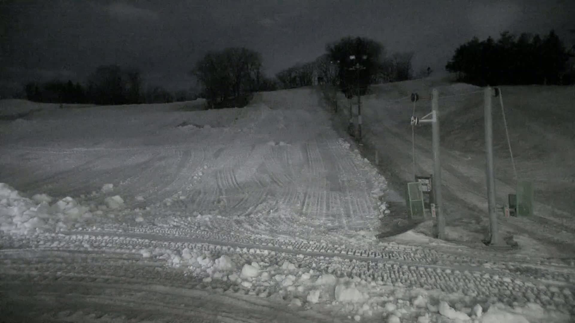 A snow-covered ski slope at night, illuminated by artificial light, features a ski lift and tire tracks in the snow.