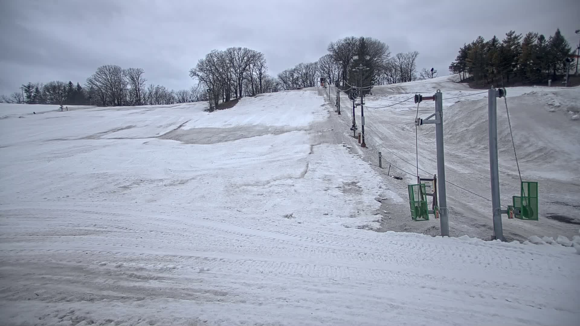 A snow-covered ski slope with a chairlift and leafless trees under a cloudy sky.