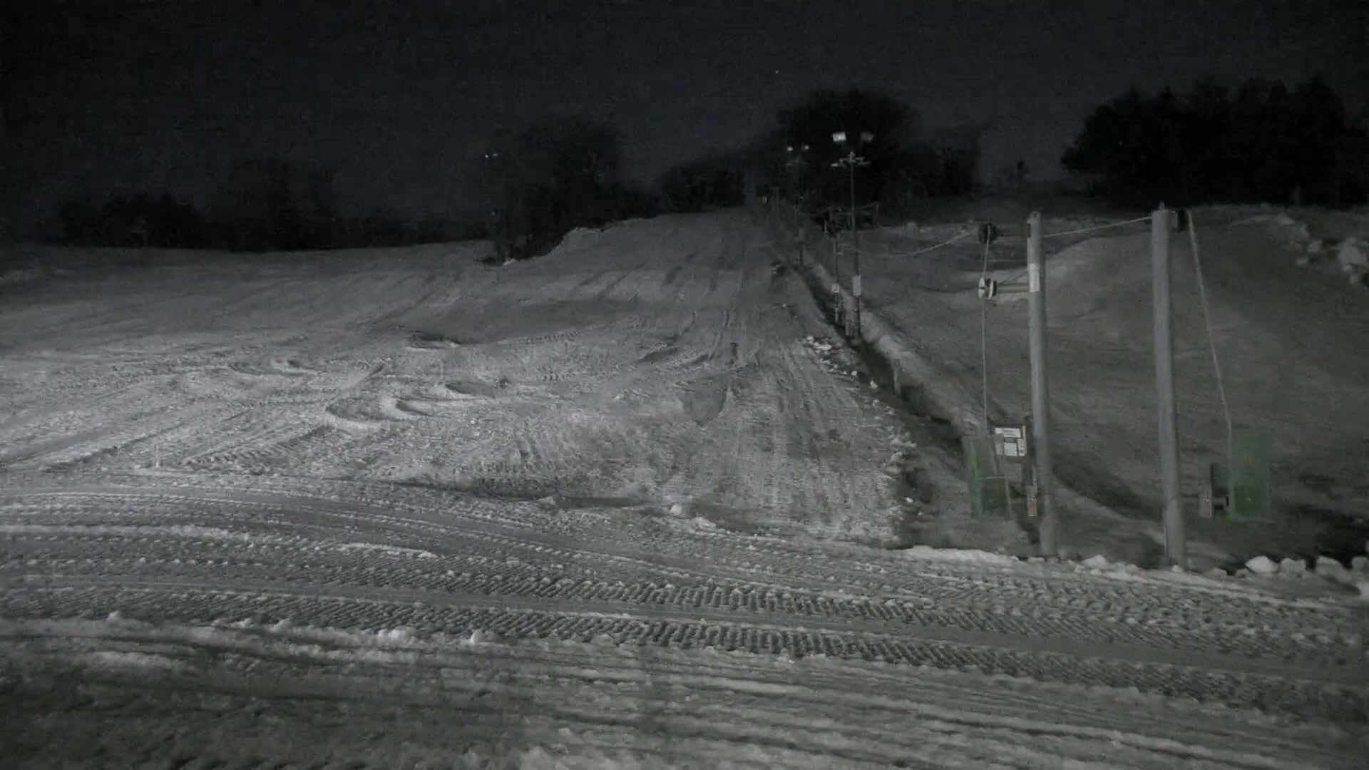 A snow-covered ski slope at night, illuminated by lights, with a ski lift visible to the right.