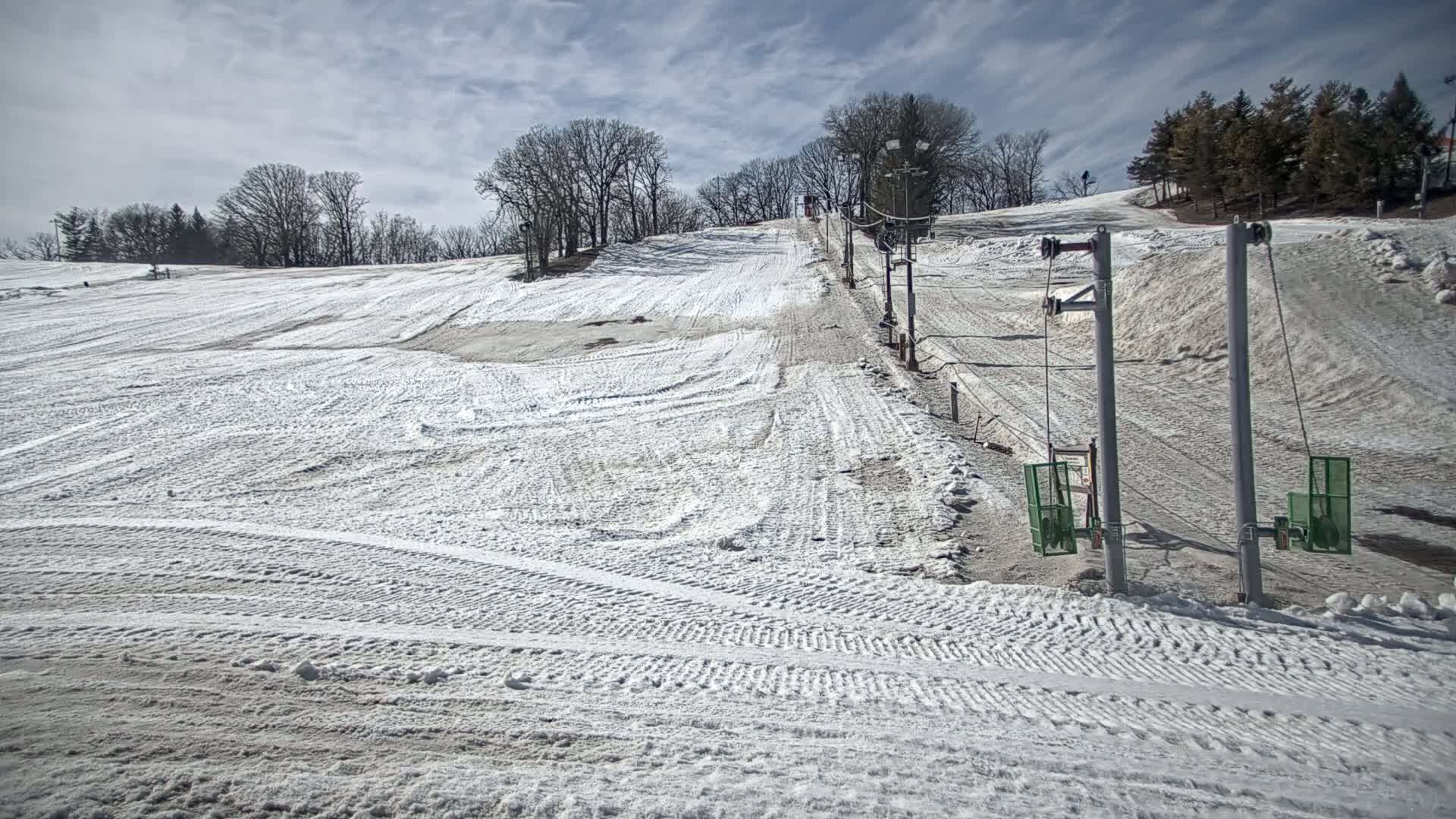 A snow-covered ski slope with a sparsely used surface lift under a partly cloudy sky.