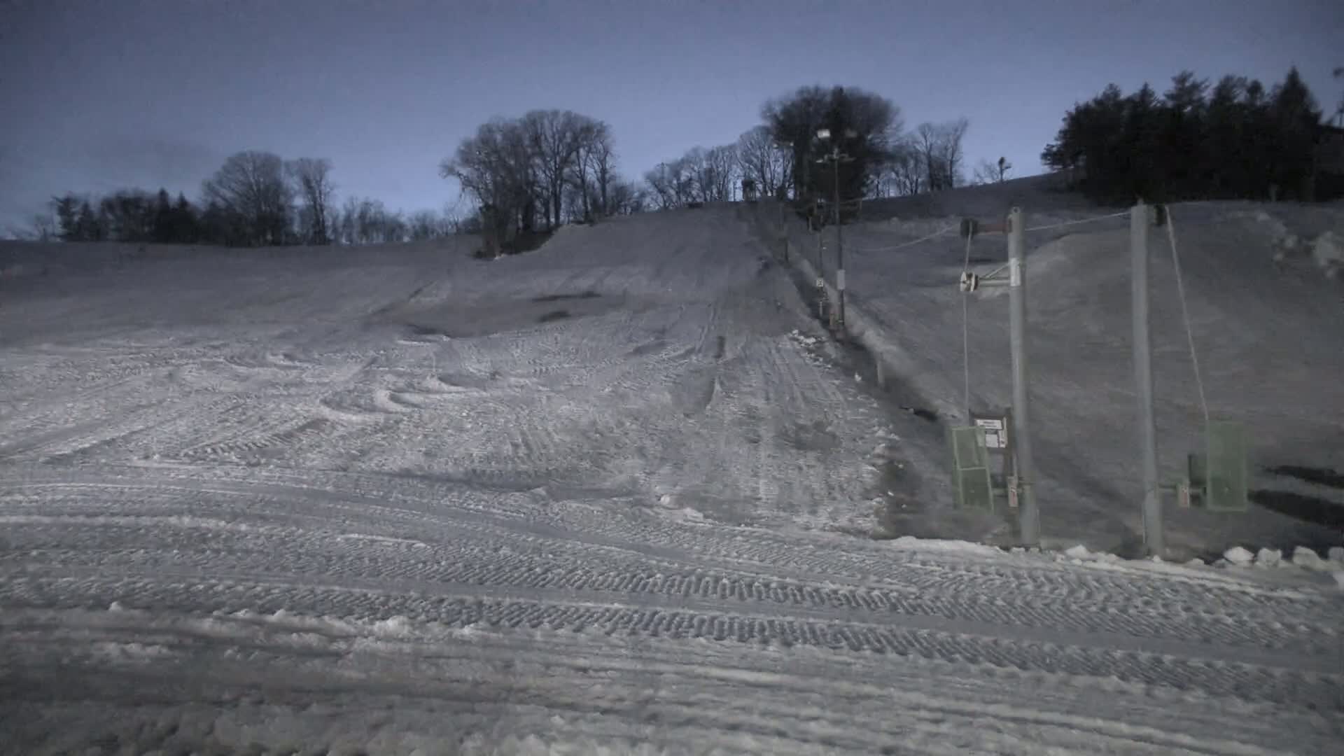 A snow-covered ski slope at dusk, with a ski lift and trees in the background.