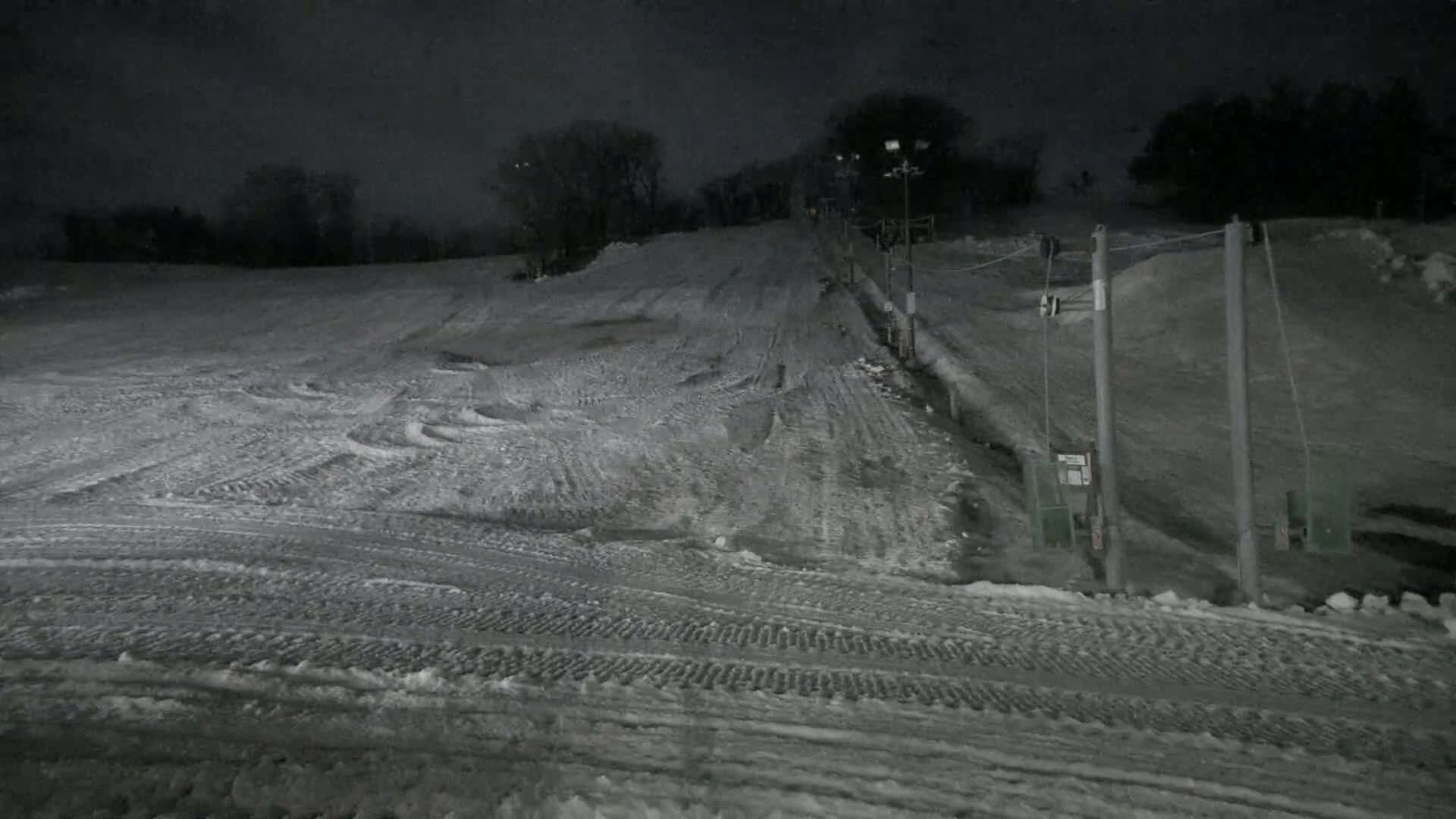 A snow-covered ski slope at night, illuminated by artificial lights, shows tire tracks and the infrastructure of a ski lift.