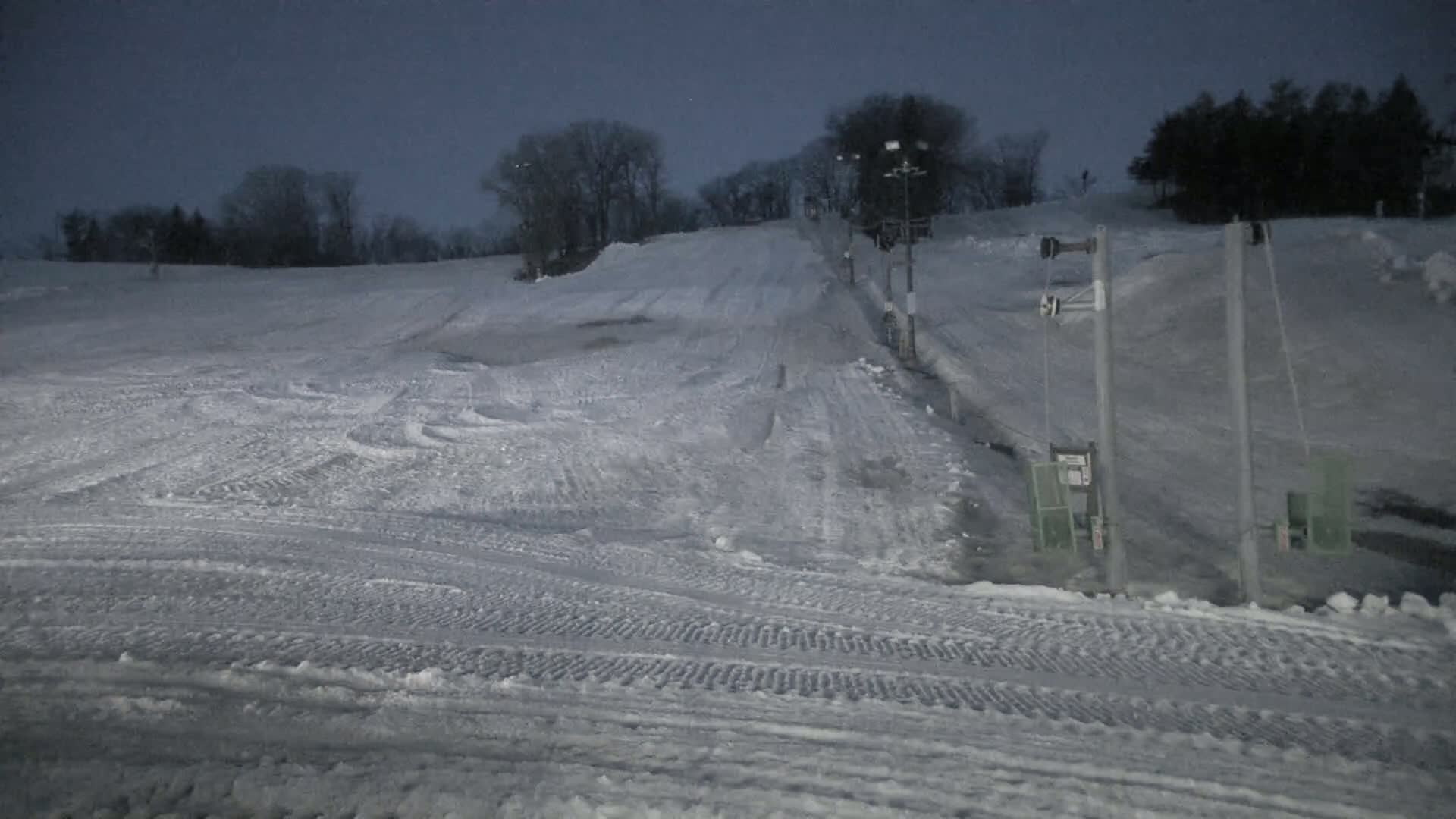 A snow-covered ski slope at night, illuminated by dim lighting, features a chairlift and trees in the background under a dark sky.