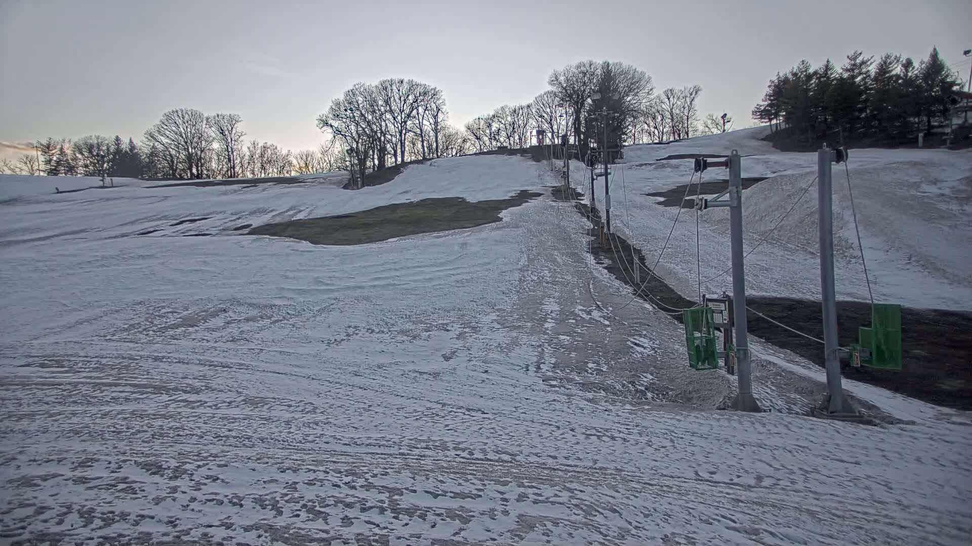 A snow-covered ski slope with a surface lift in the late afternoon or early evening under a cloudy sky.