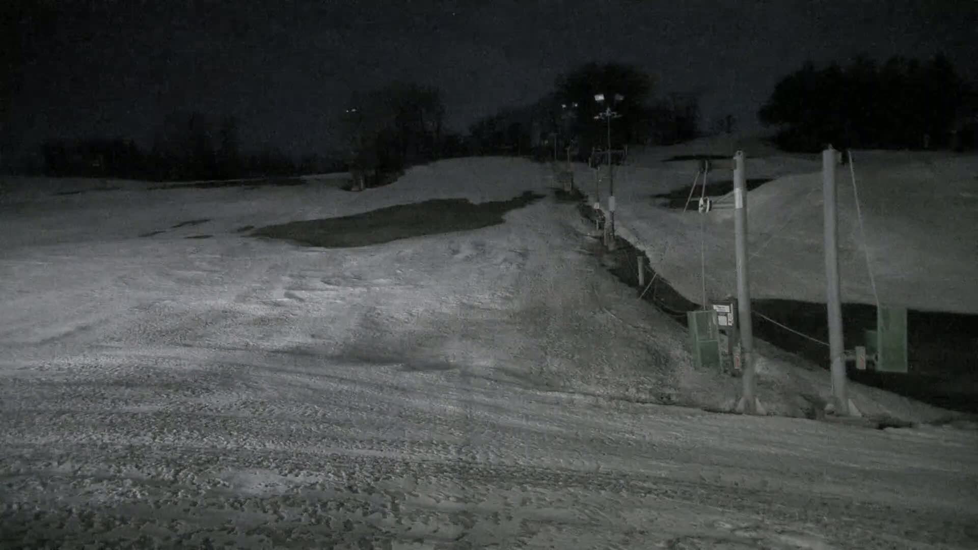 A snow-covered ski slope at night, illuminated by lights, with a ski lift visible in the lower right.