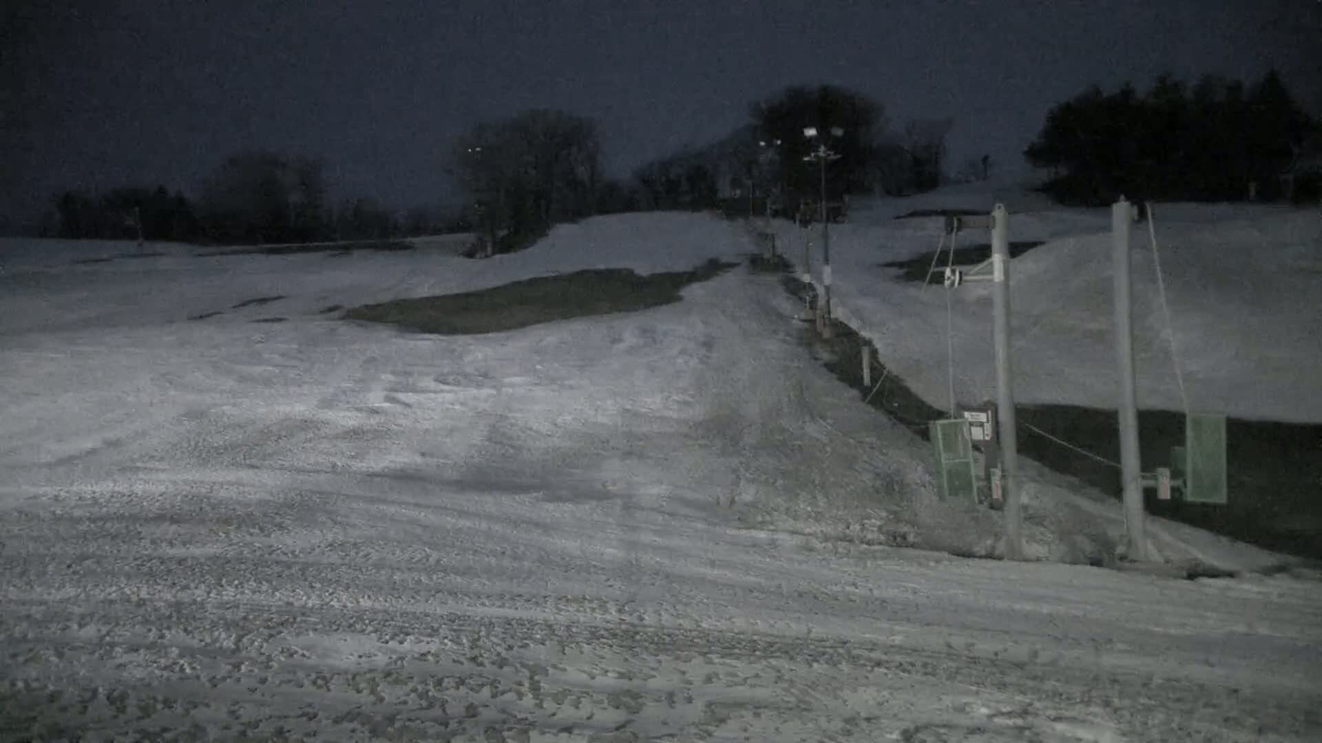 A snow-covered ski slope at night, illuminated by artificial lights, with a ski lift in the foreground.
