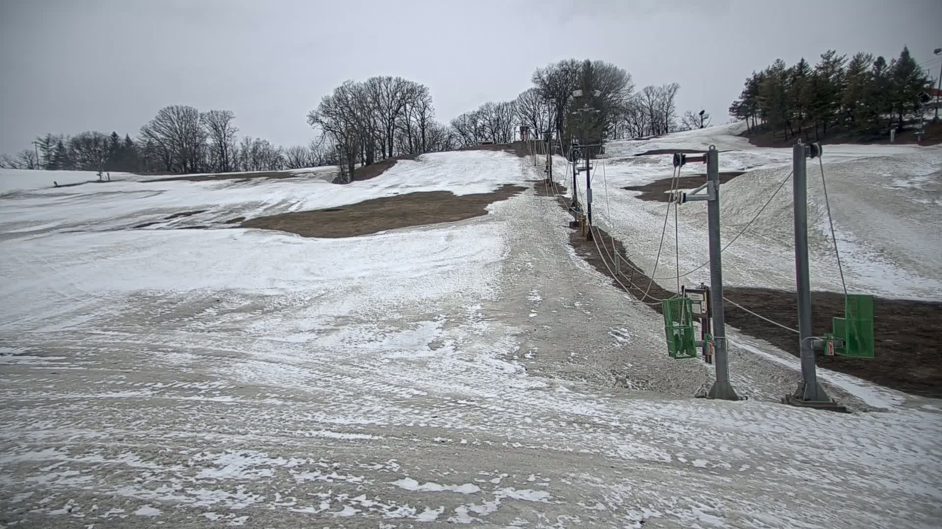 A sparsely snow-covered ski slope with a chairlift under an overcast sky.
