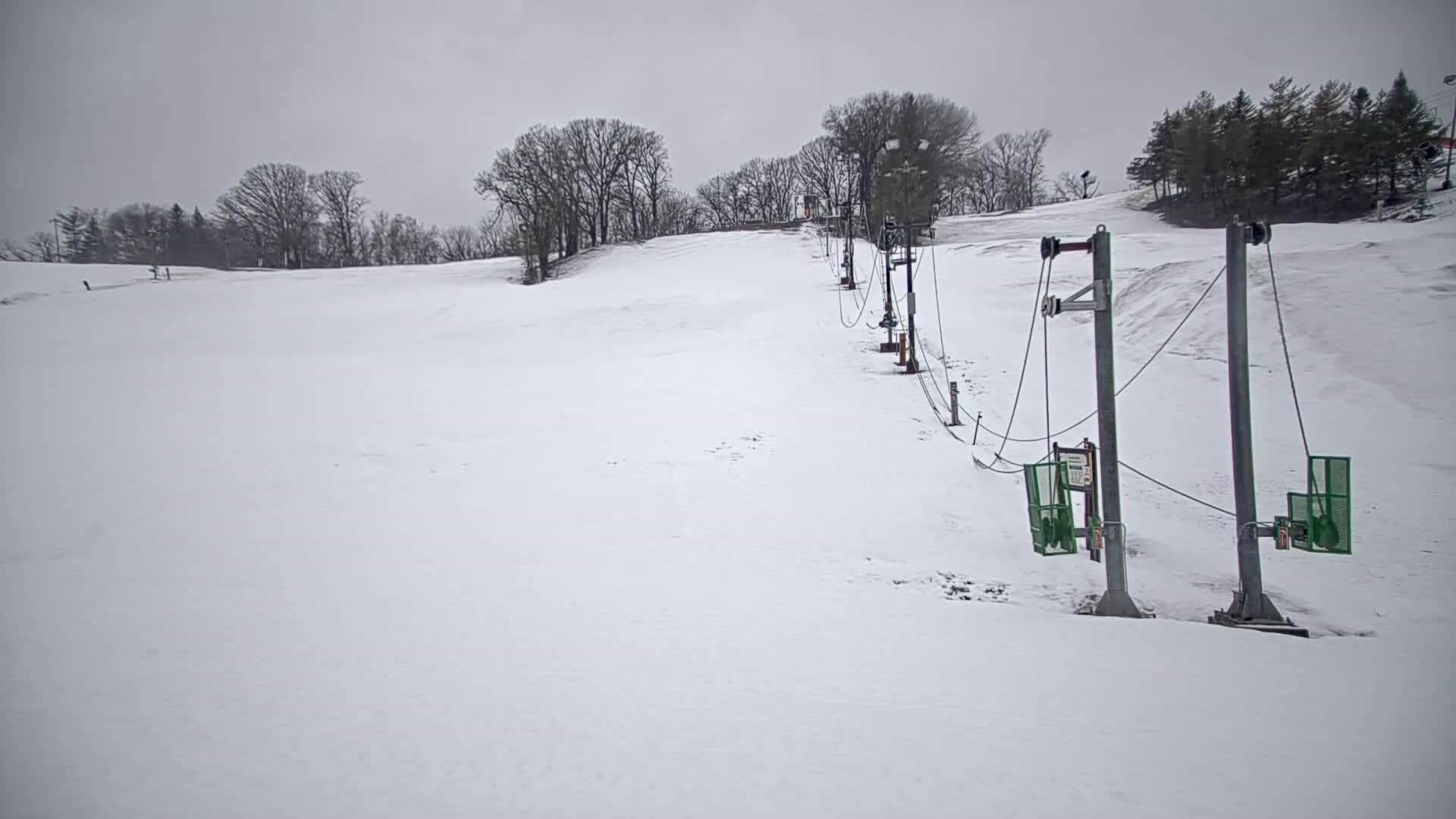 A snow-covered ski slope with a surface lift in the foreground under an overcast sky.