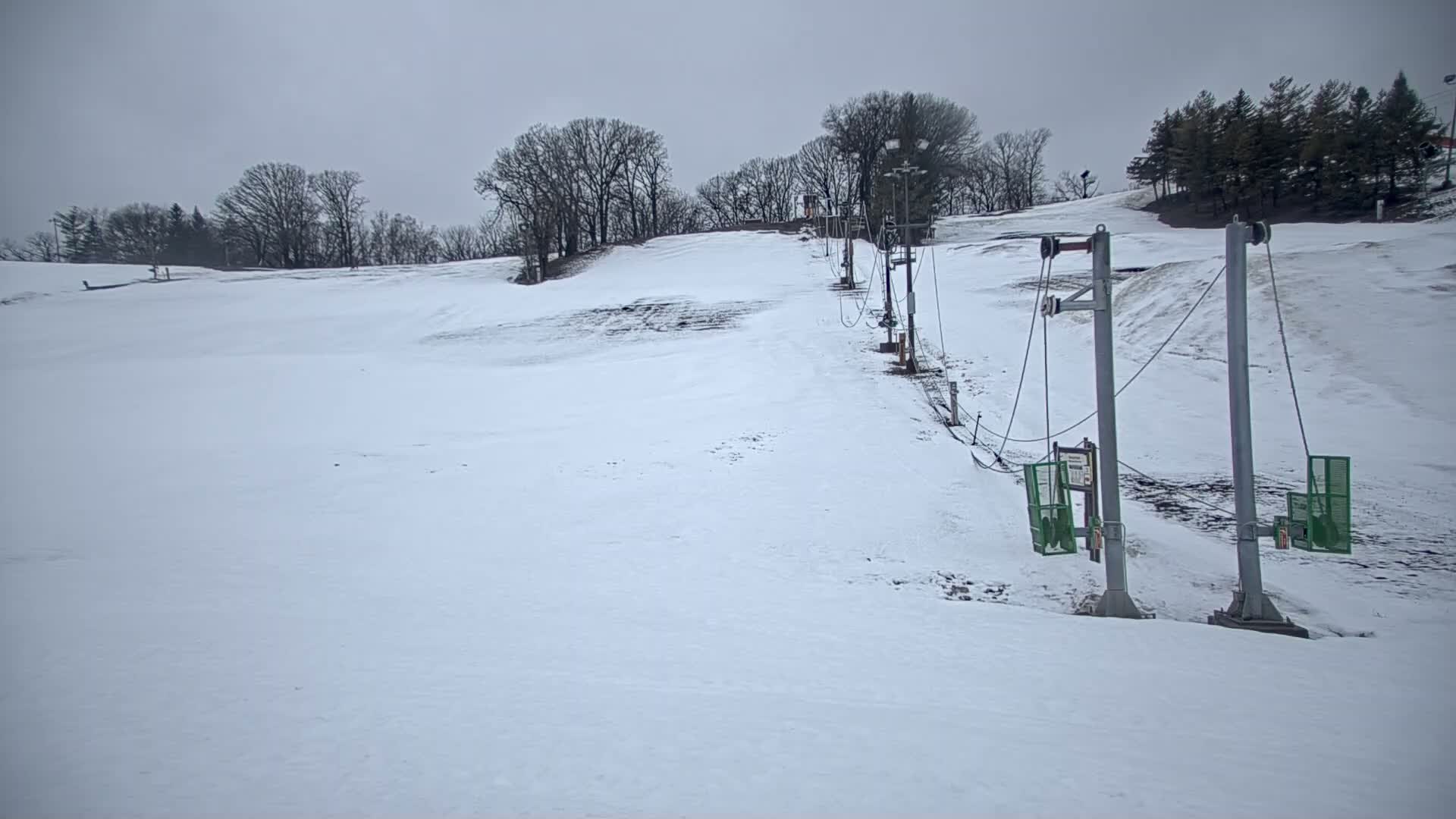 A snow-covered ski slope with a sparsely used surface lift ascends a hill under an overcast sky.