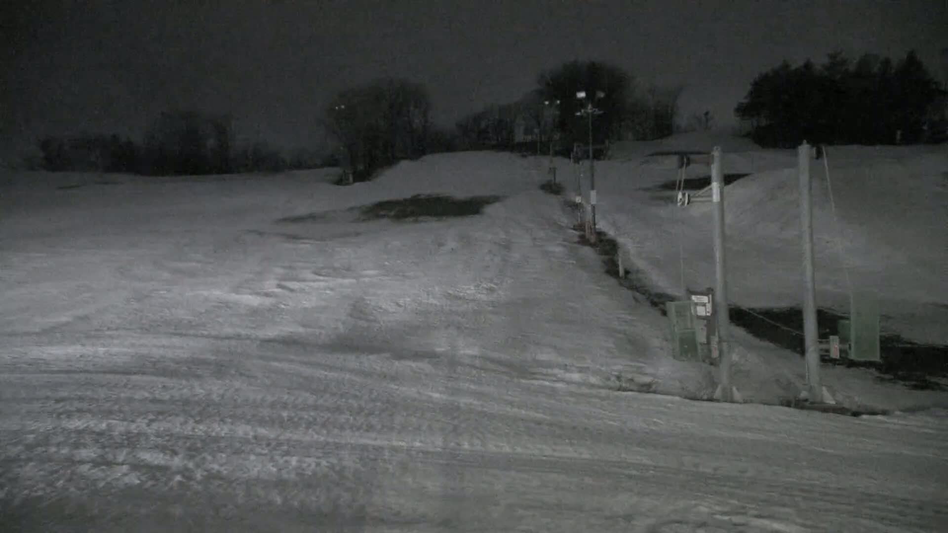 A snow-covered ski slope at night, illuminated by lights, with a ski lift visible in the foreground.