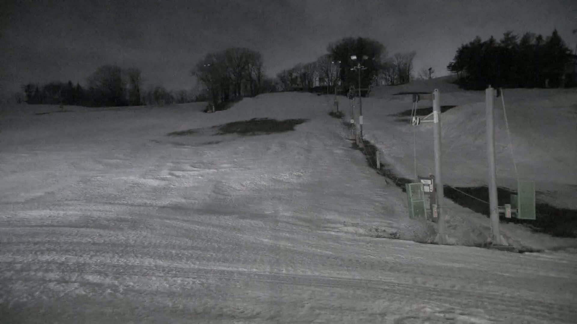 A snow-covered ski slope at night, illuminated by artificial lights, with a sparsely used ski lift visible in the foreground.