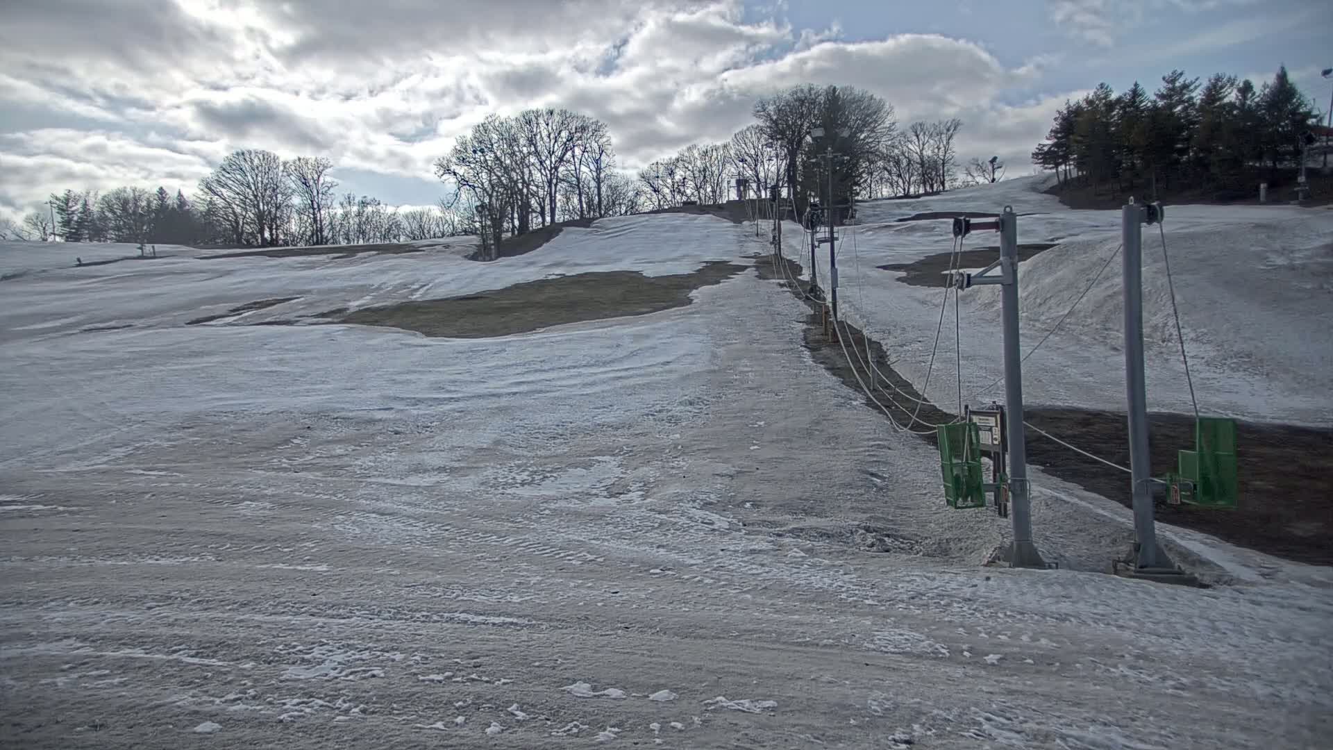 A sparsely snow-covered ski slope with a chairlift under a partly cloudy sky.