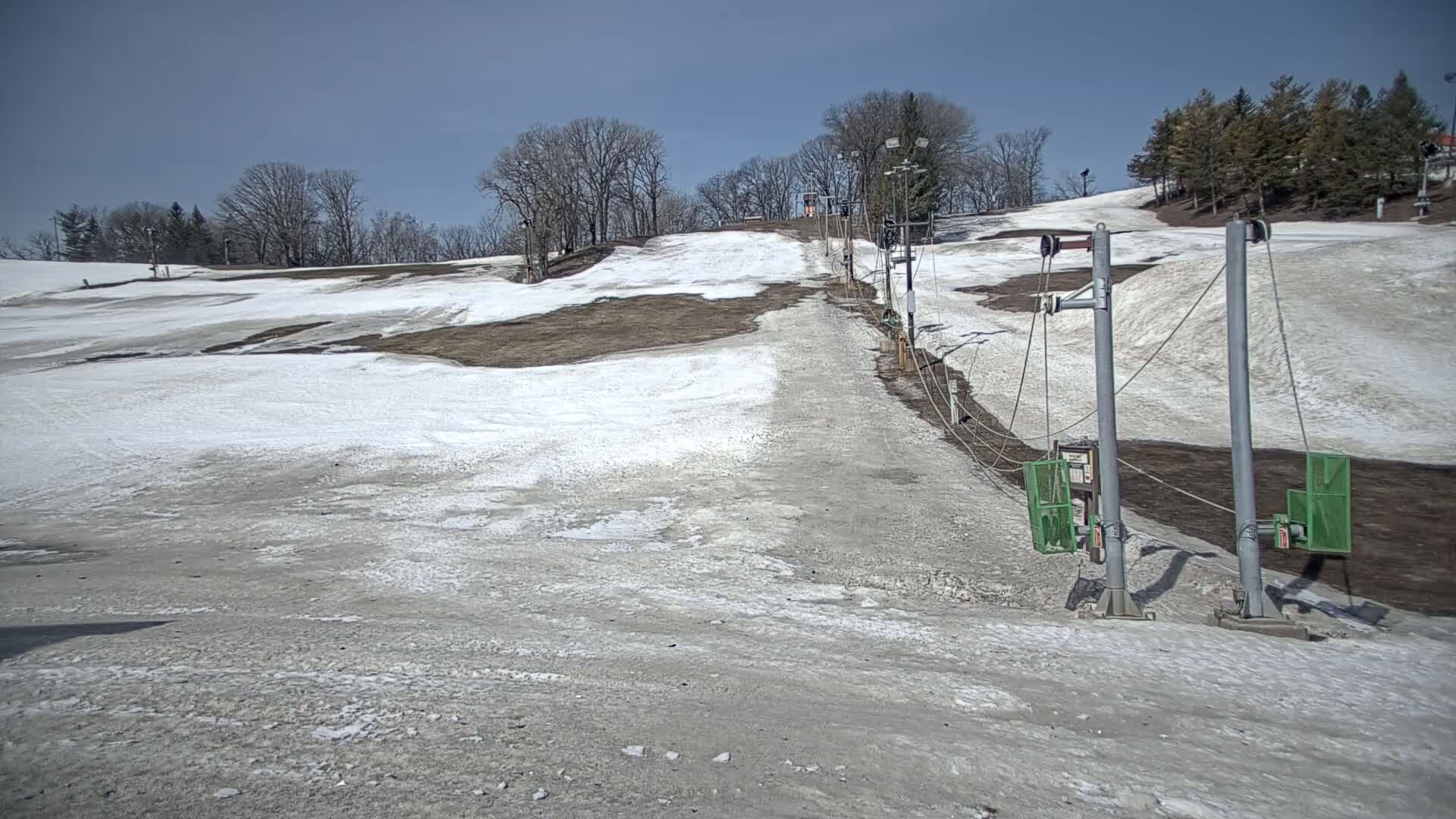 A mostly bare ski slope with patches of snow and a surface lift under a clear blue sky.