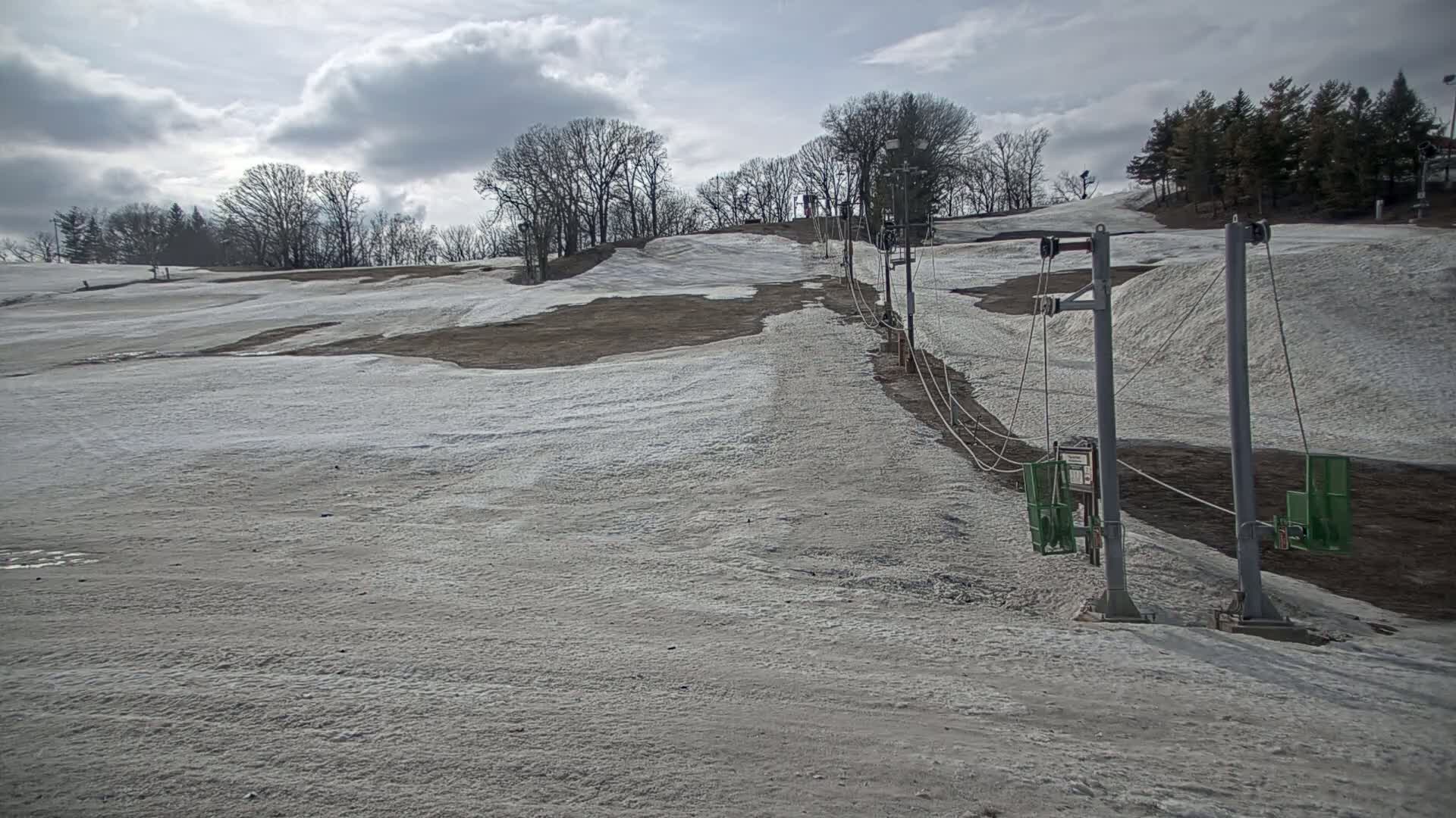 A sparsely snow-covered ski slope with a chairlift under a cloudy sky.