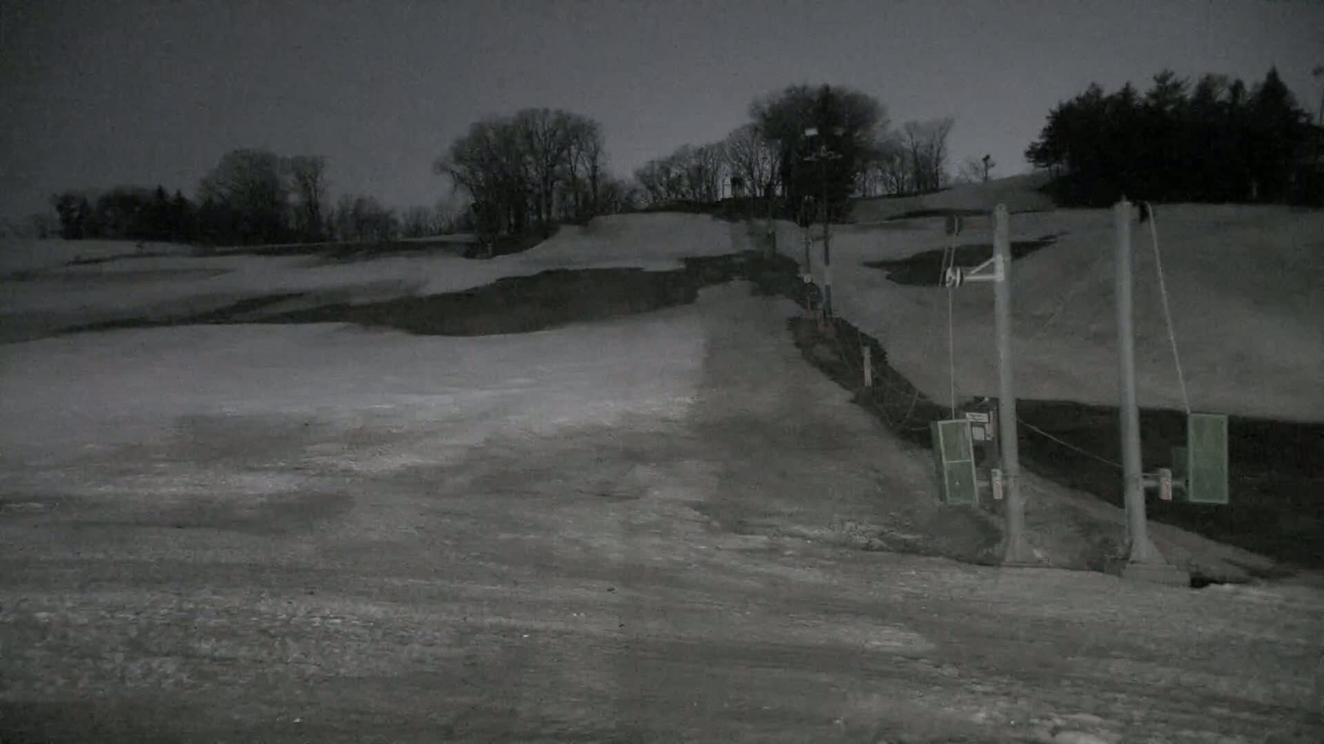 A snow-covered ski slope at night, illuminated by artificial light, with a ski lift visible in the foreground.