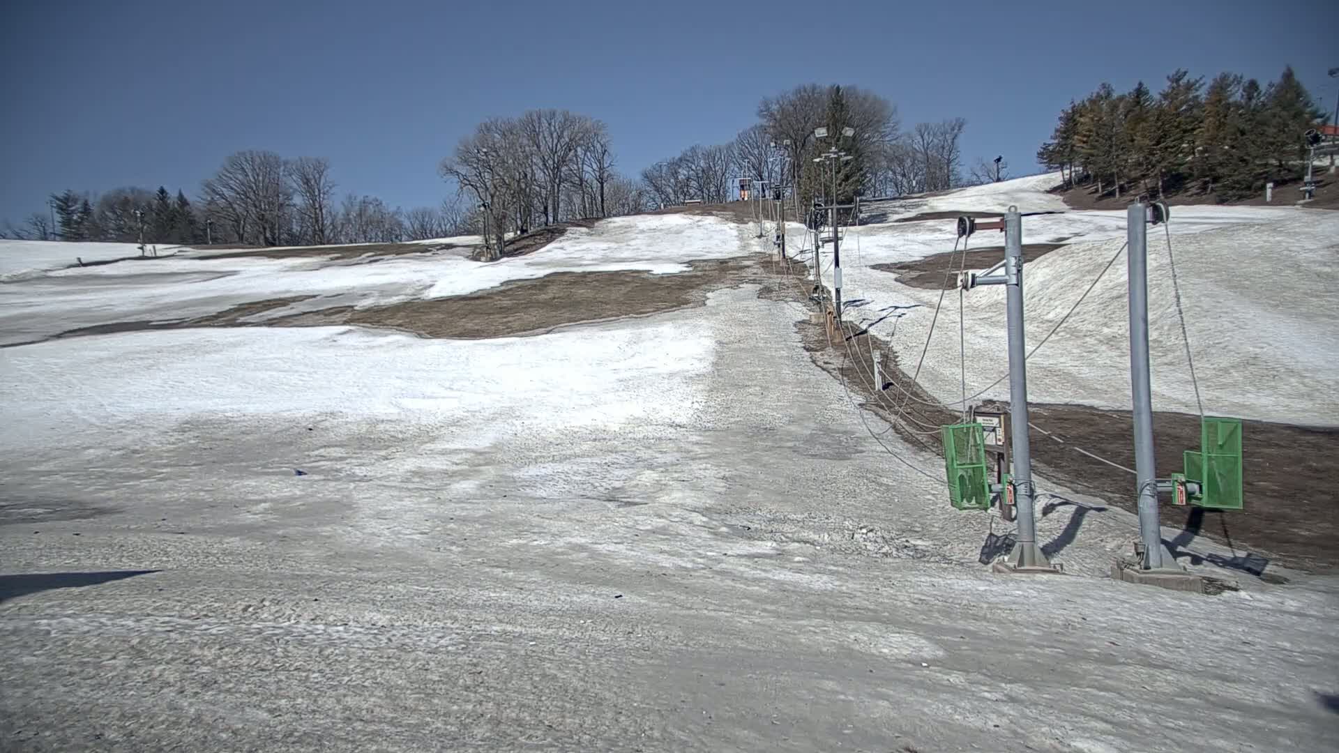 A ski slope with patchy snow cover and a chairlift under a clear blue sky.