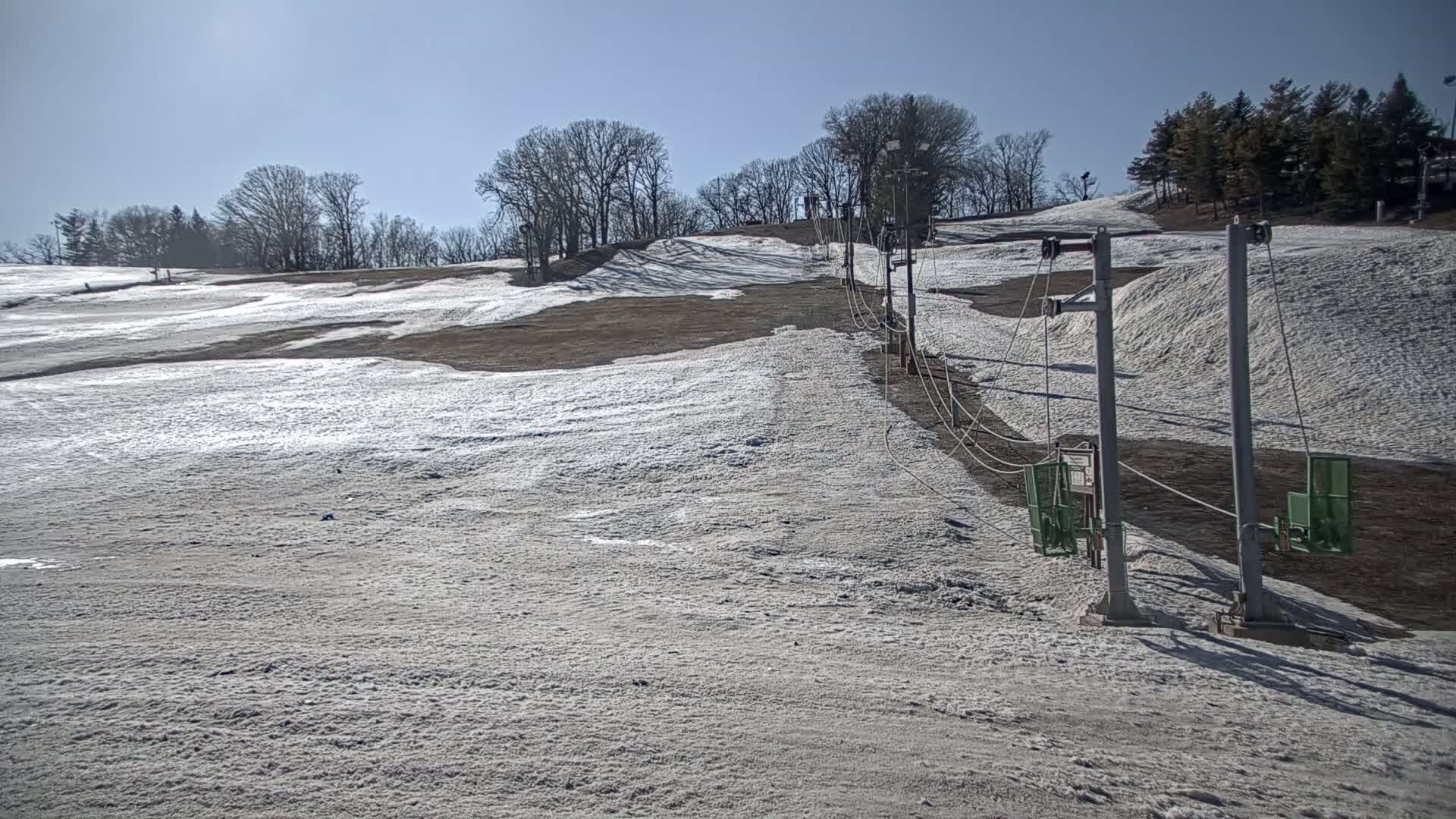 A ski lift stands on a sparsely snow-covered hill under a clear, sunny sky.