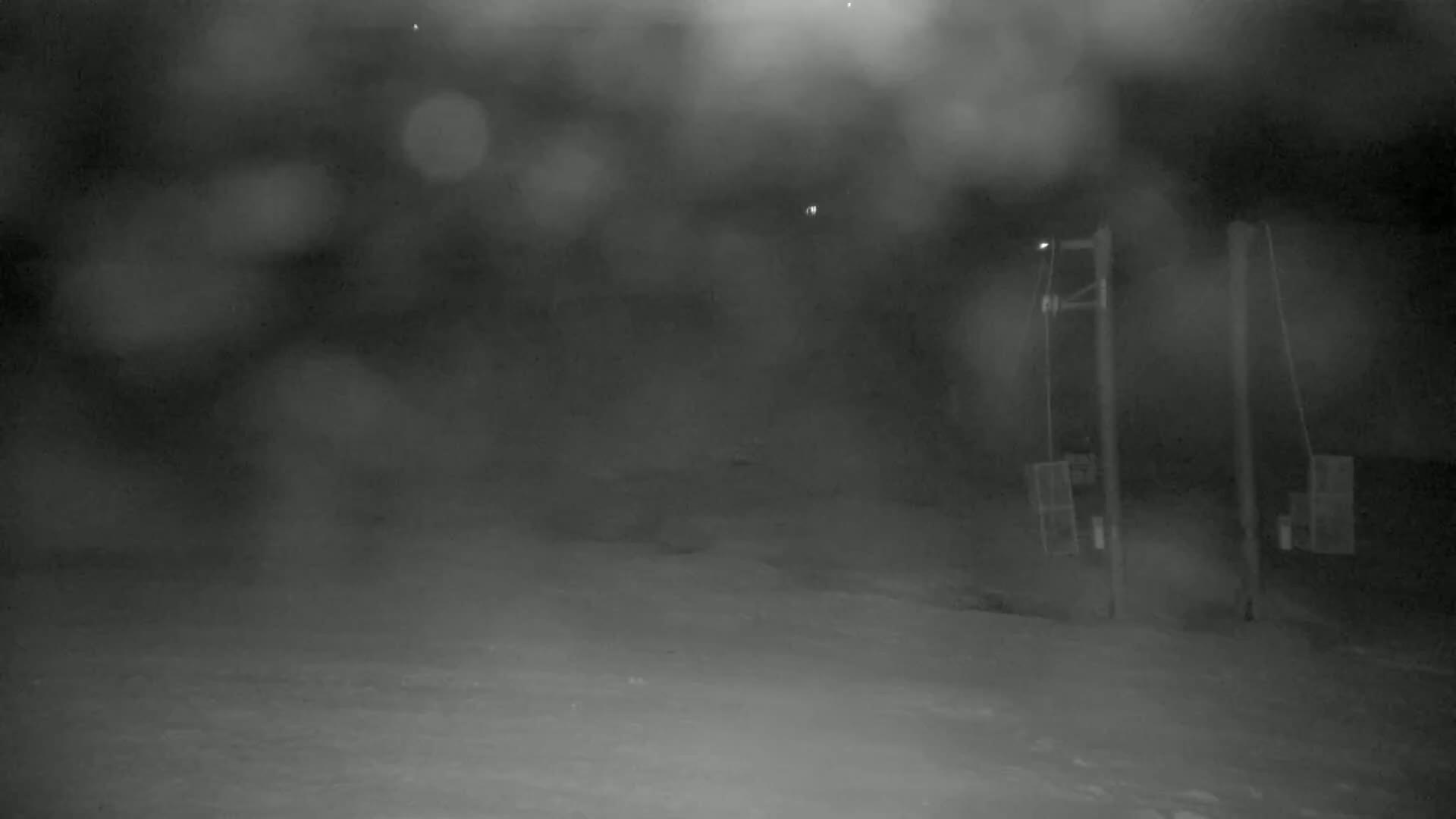 A snowy, nighttime scene shows two utility poles with equipment attached, partially obscured by blowing snow or fog.