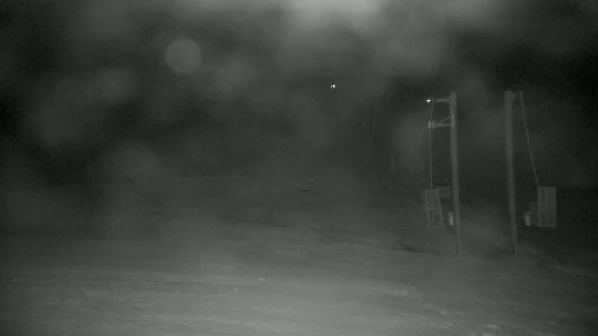 A nighttime, snowy scene shows two empty chairlift stations and a hazy, blurry background.