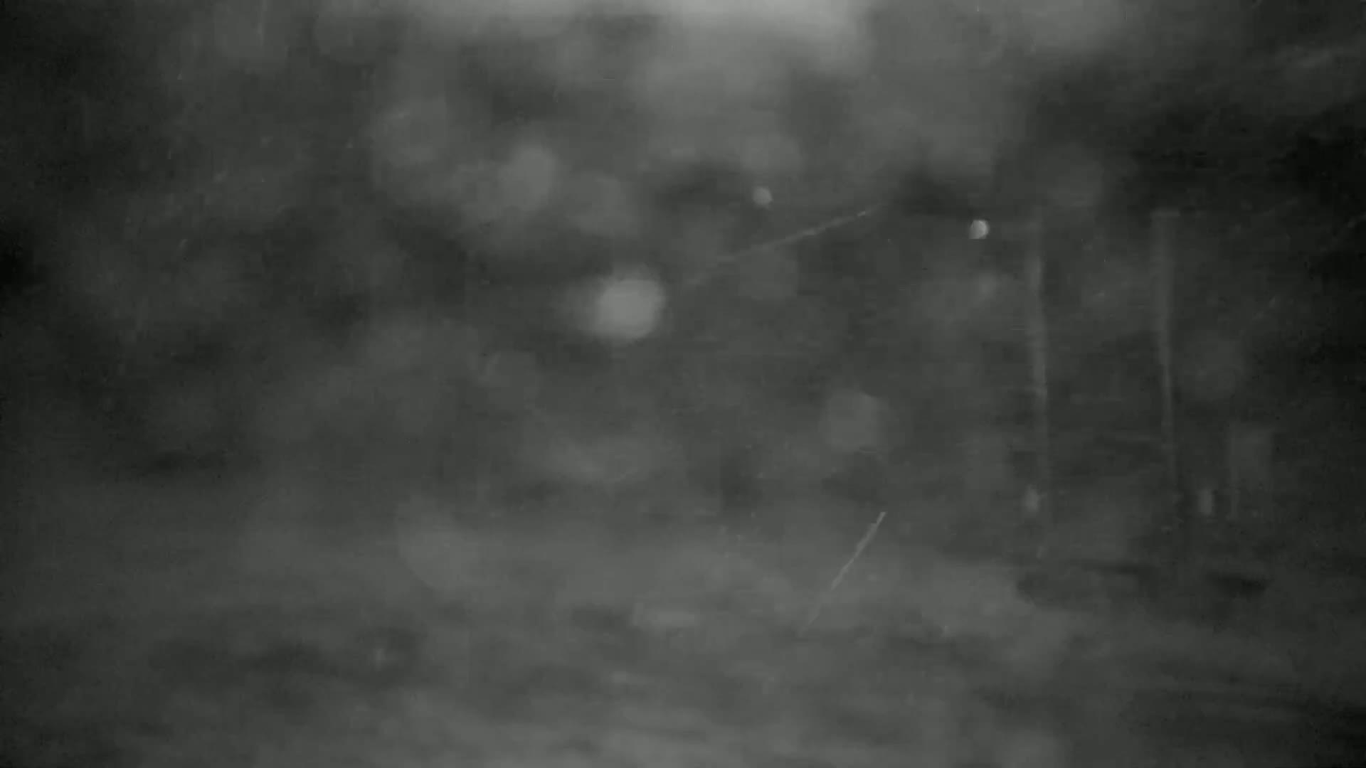A blurry, dark, and snowy outdoor scene shows indistinct shapes that may be buildings or trees.