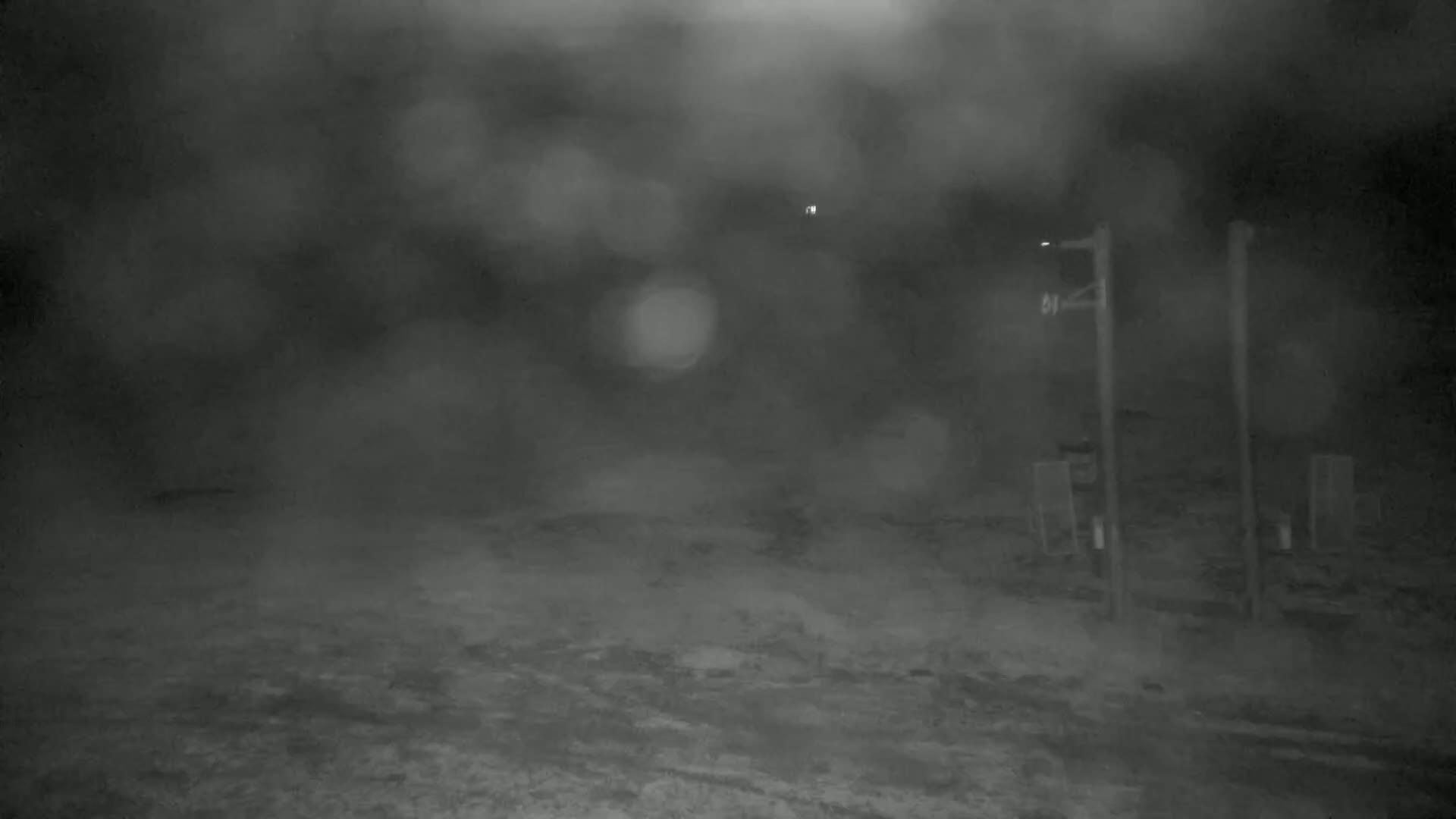 A nighttime, blurry outdoor scene shows a flat, snow-covered ground with two tall posts and indistinct shapes in the distance under a heavy, foggy sky.