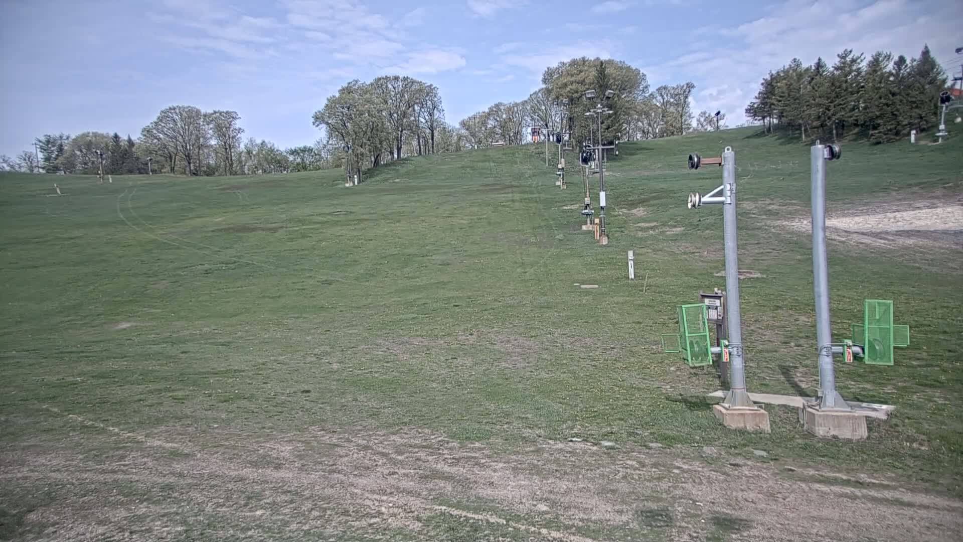 A grassy hill with a ski lift and trees under a partly cloudy sky.