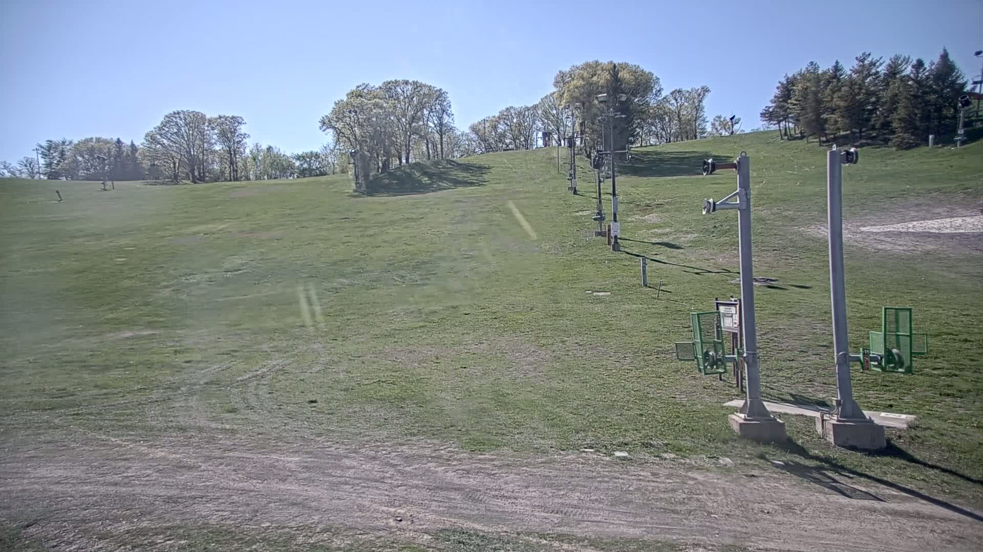 A grassy hill with a ski lift and several deciduous trees under a clear, sunny sky.