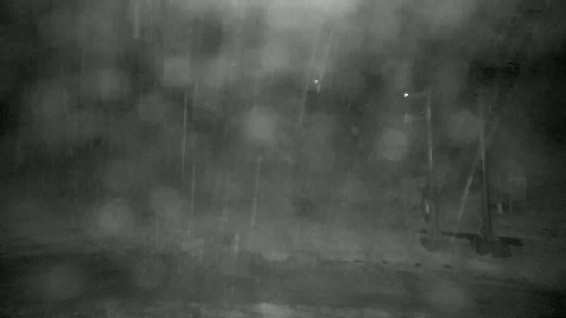 A blurry, dark image shows heavy rain falling at night, obscuring the view of indistinct structures and objects in the distance.