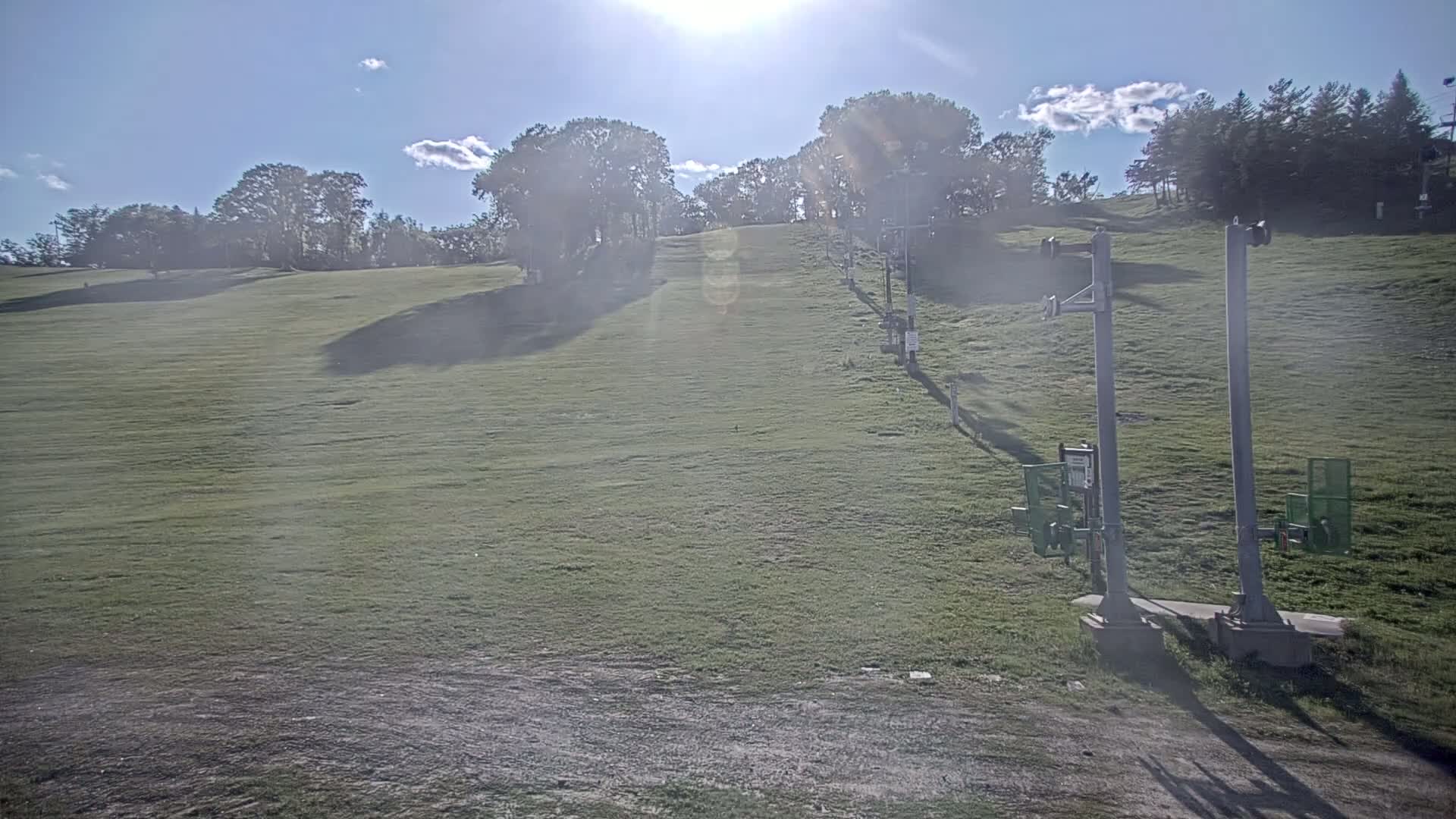 A grassy hill with a ski lift in the foreground under a mostly sunny sky.