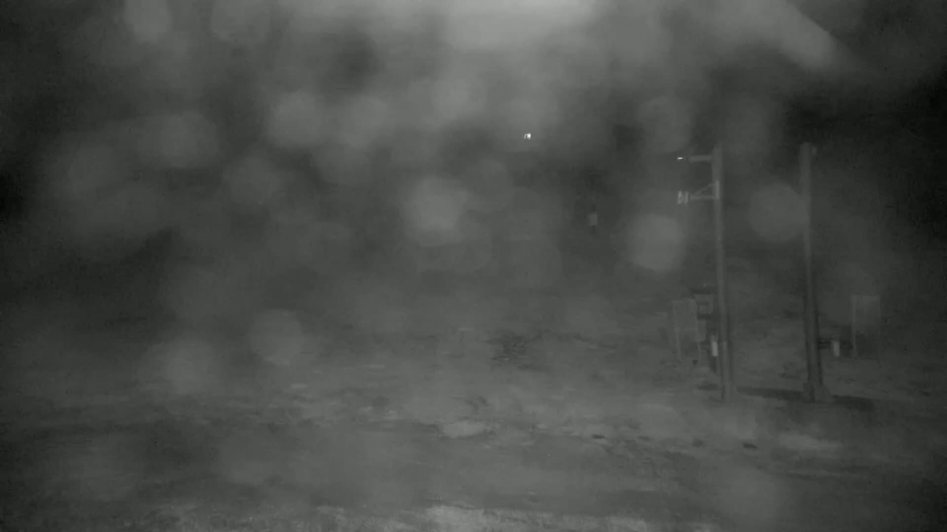 A blurry, nighttime image shows a sparsely populated area with some poles and indistinct objects in the distance, under heavy rain or fog.